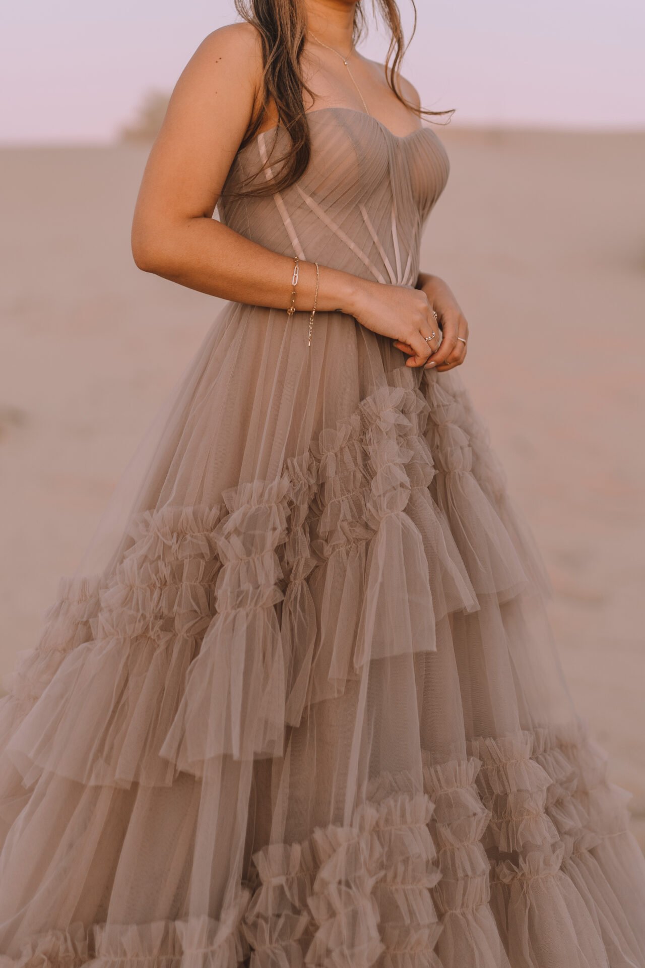 A woman wearing a beige strapless layered tulle dress standing outdoors with blurred background.