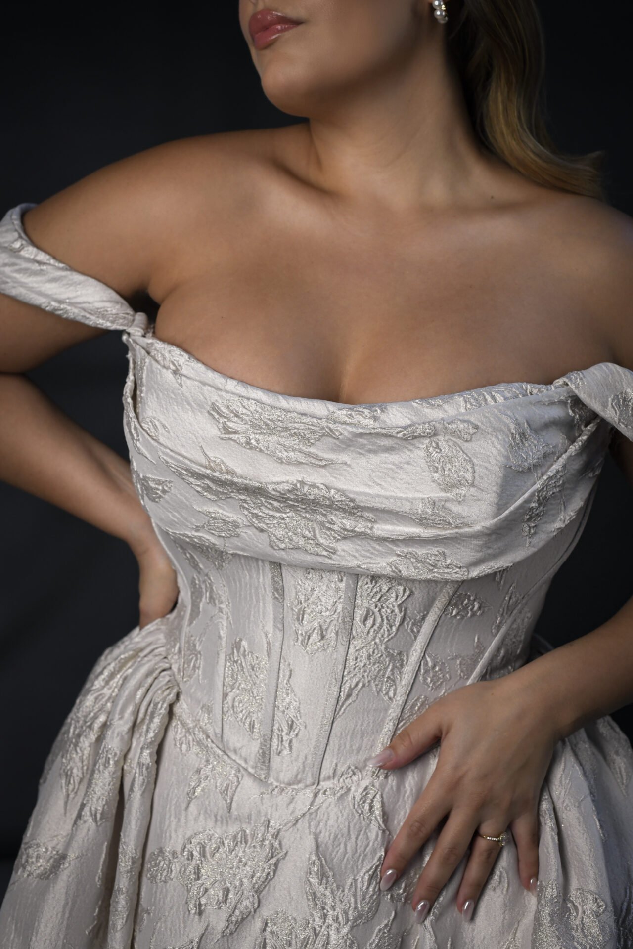 A woman wearing a strapless, cream-colored dress with floral patterns, showing her neck, shoulders, and part of her face. She is standing against a dark background, with one hand on her hip and the other resting on her waist.
