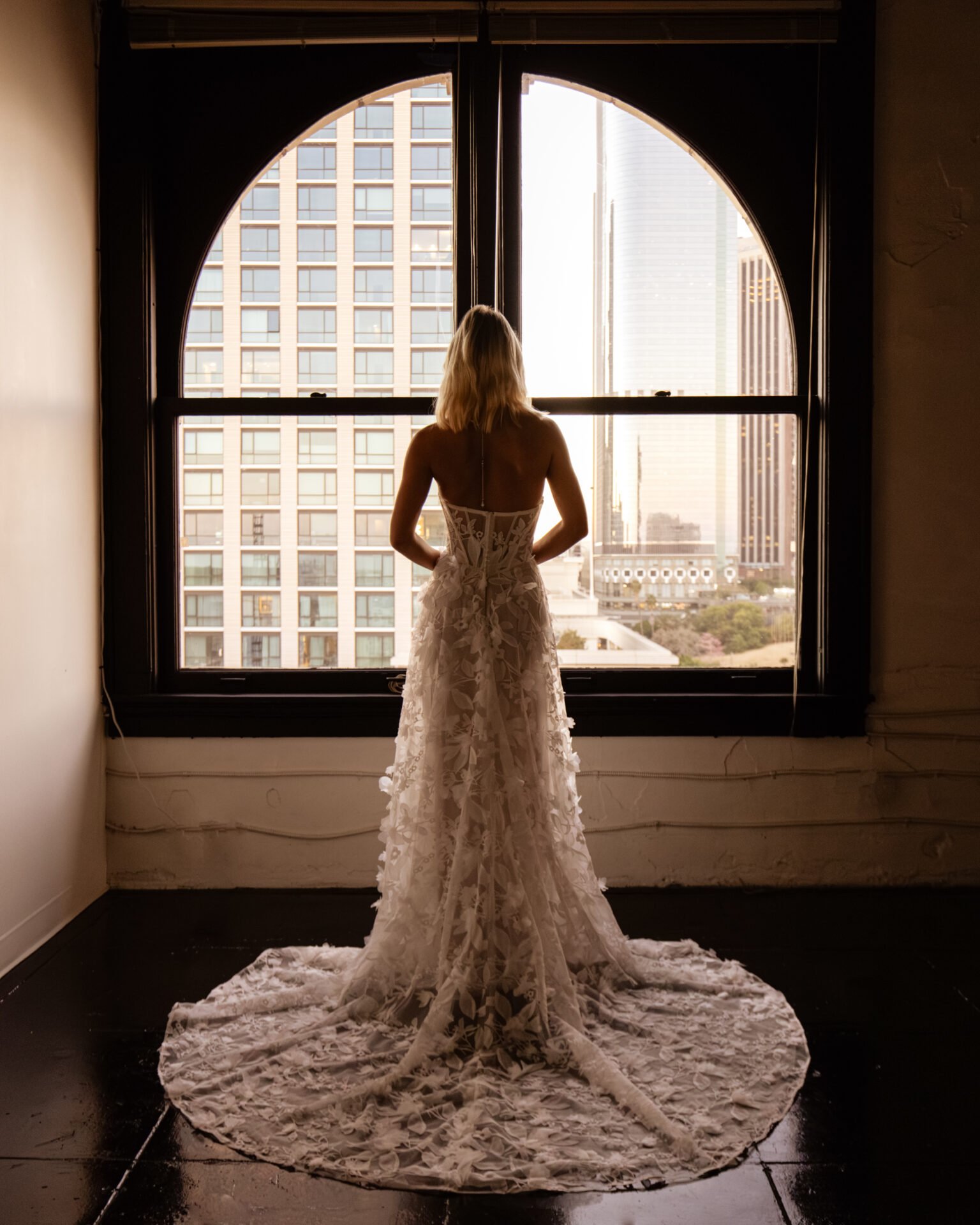 A woman in an elegant wedding gown standing in front of a large window, overlooking a cityscape with tall buildings, during sunset.