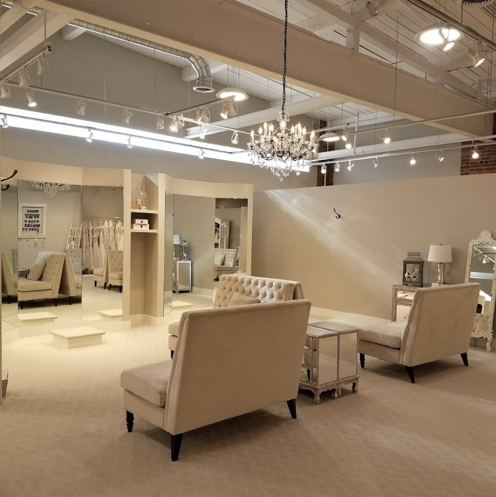 A furniture showroom with beige upholstered chairs and sofas, a chandelier, a large mirror, and display tables, in a bright, modern space.