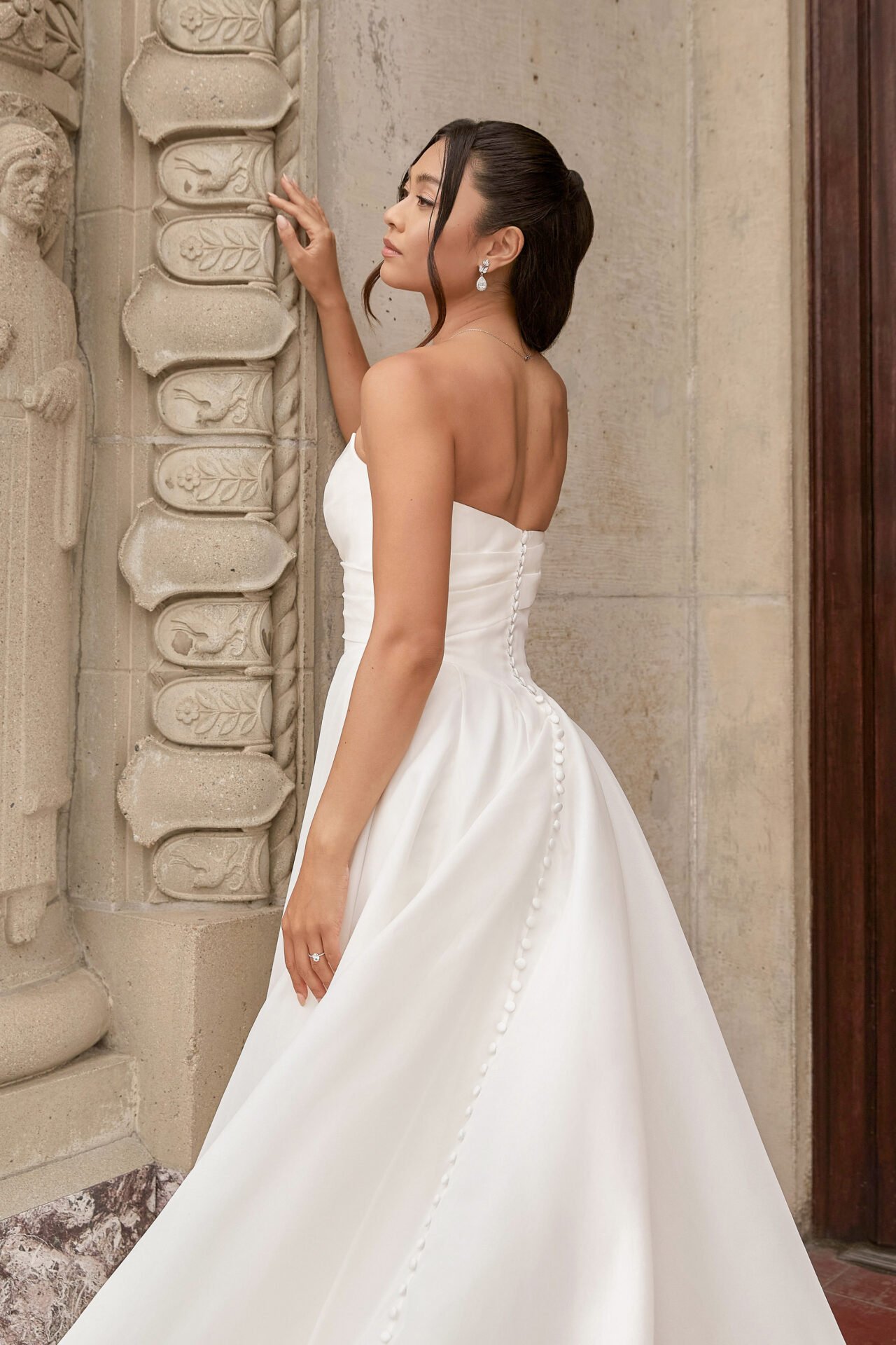 A woman in a white strapless wedding gown with buttons down the back, standing against a stone wall.