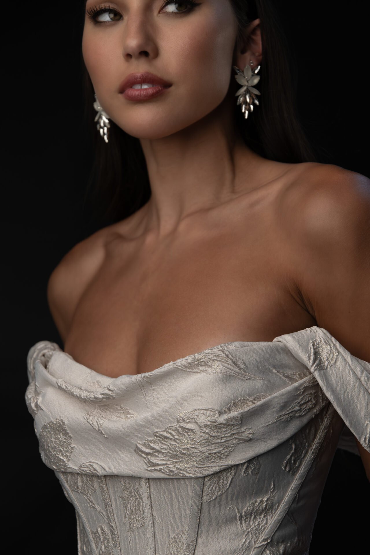 Close-up of a woman with dark hair wearing floral earrings and a strapless, textured satin dress against a black background.