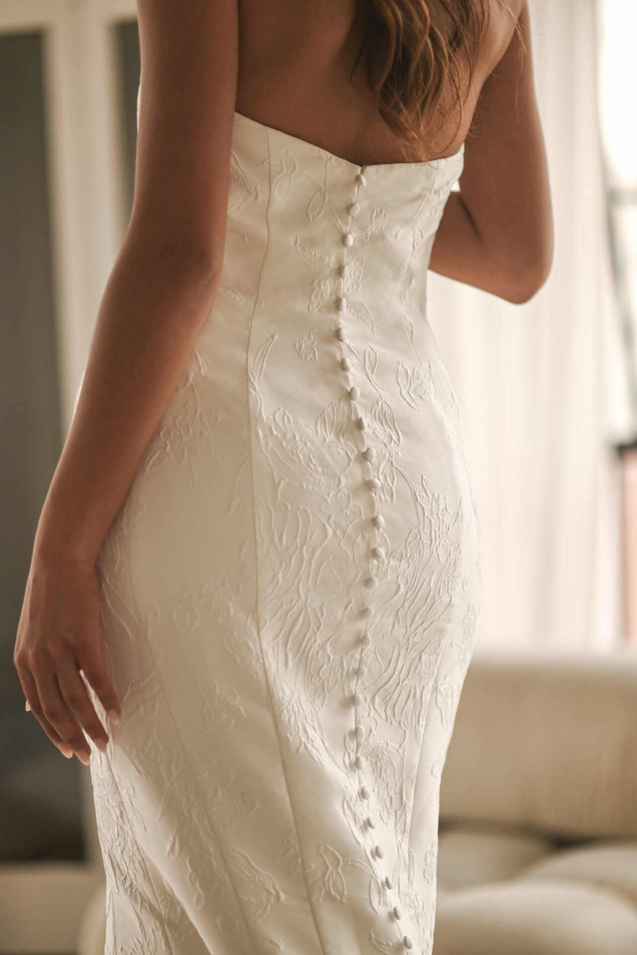 Close-up of a woman's back in a white, textured wedding dress with buttons running down the center and a partial view of her arm and hair.