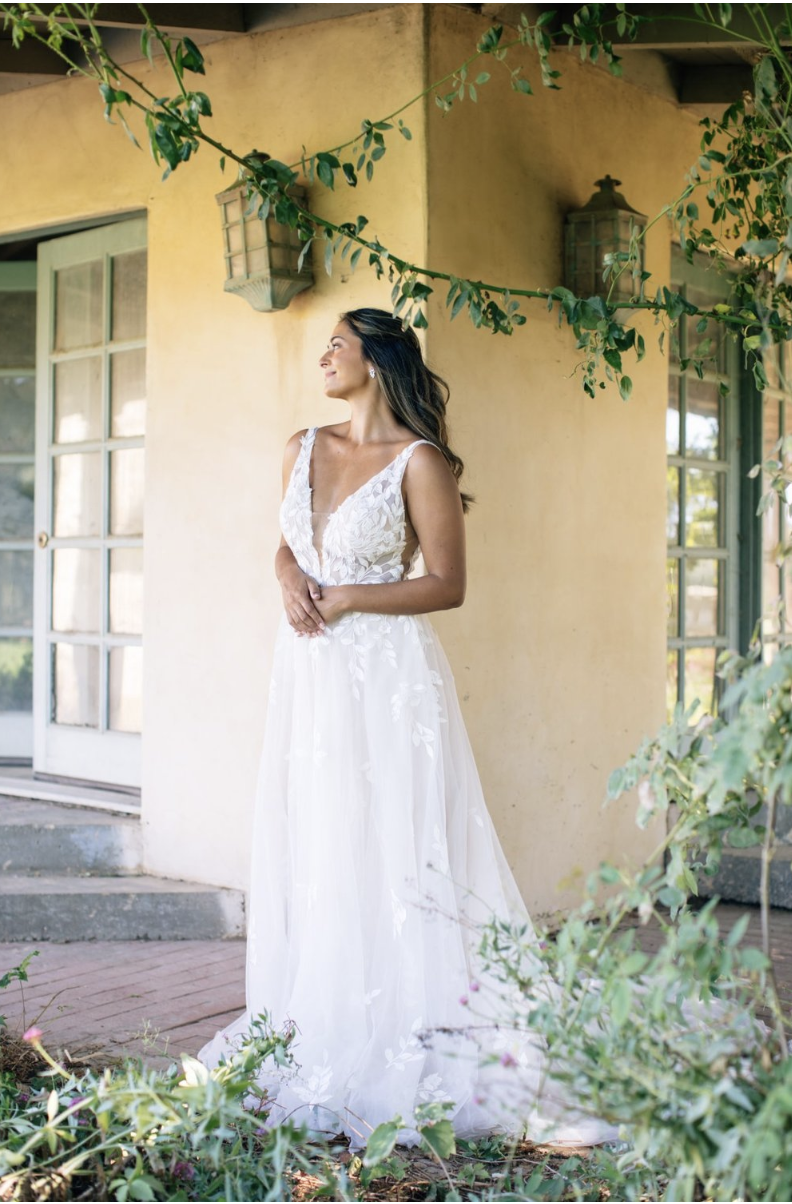 A woman in a white wedding dress standing outdoors near a beige wall, with green foliage and hanging lanterns, looking to the side.