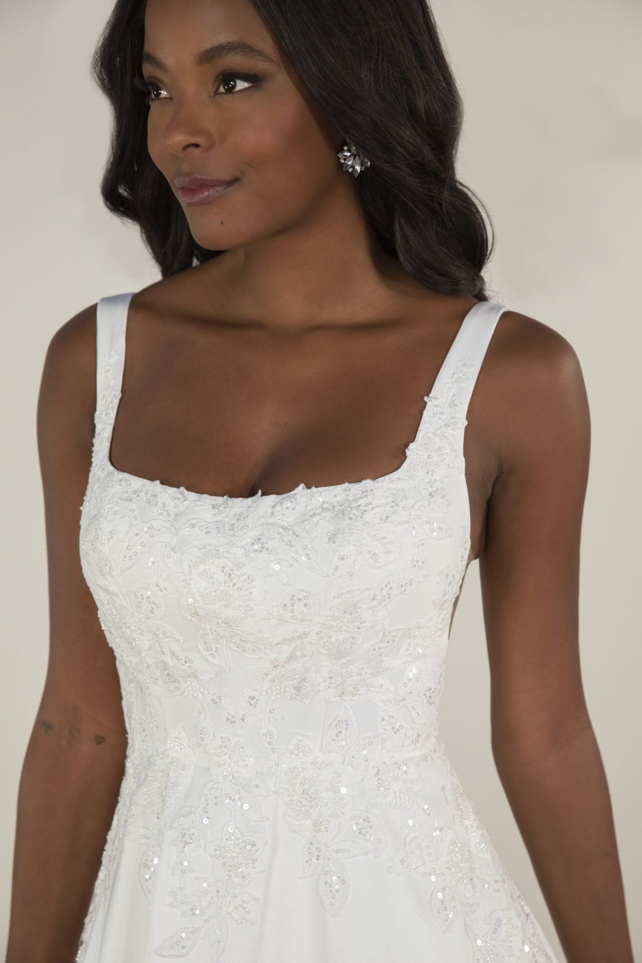 A woman wearing a white, embroidered dress with sequins and floral patterns, smiling softly, with dark hair styled in loose waves, and wearing earrings.