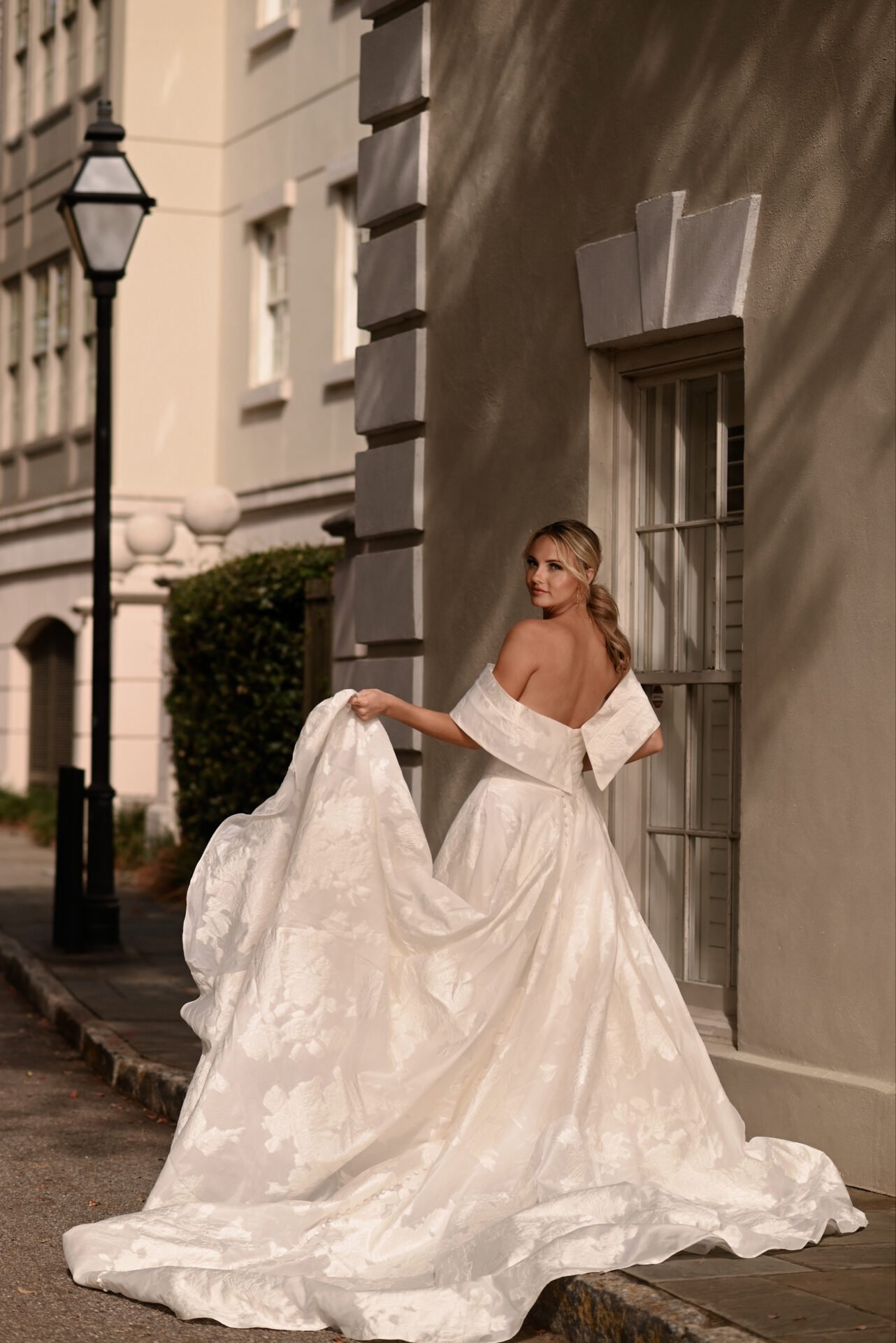 A woman in an elegant strapless wedding gown with a voluminous skirt standing on a city sidewalk at night, holding part of her dress and looking back.