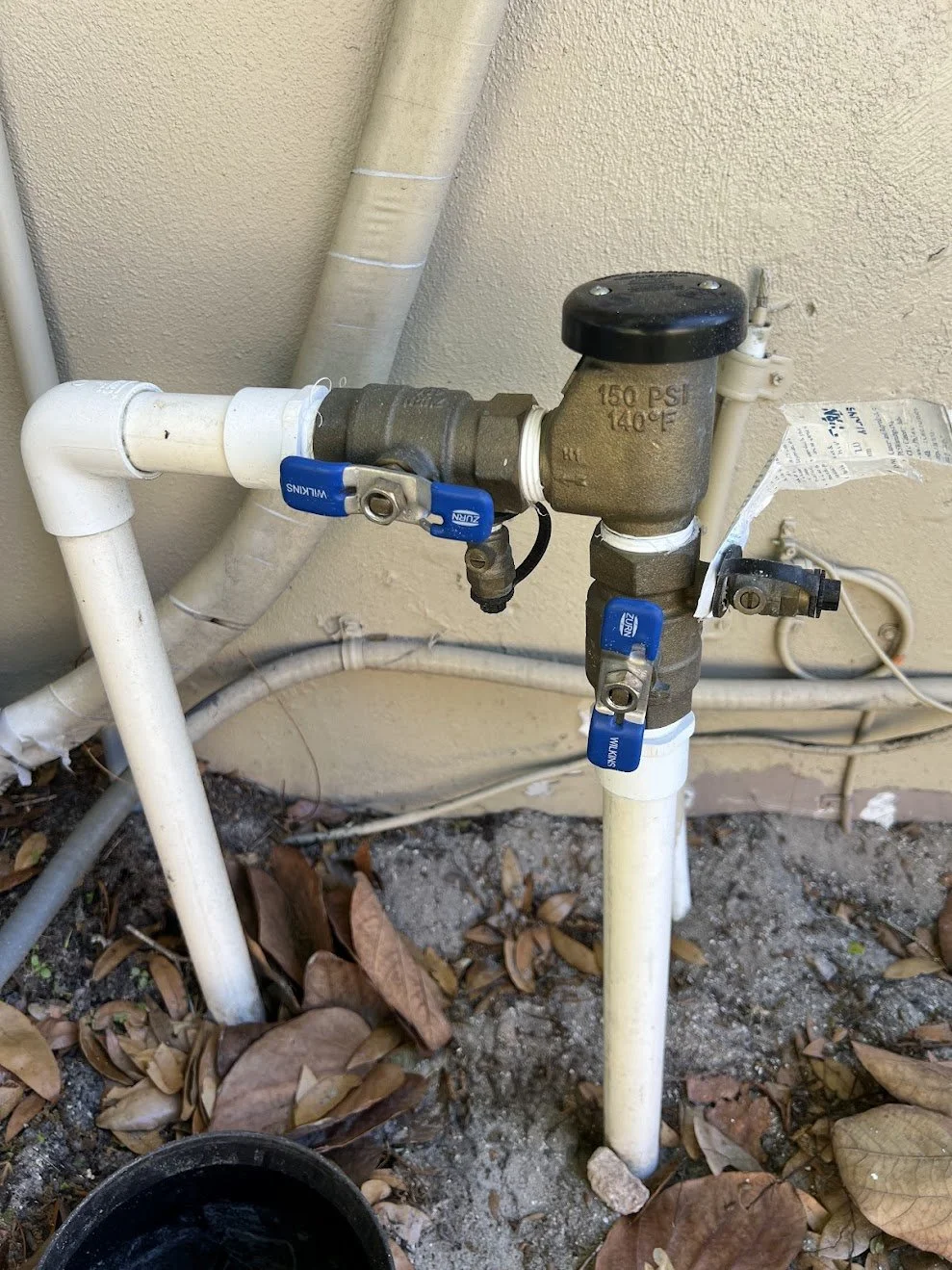Outdoor plumbing setup with white PVC pipes, a brass valve with blue handle, and a black cap, installed against a beige wall with fallen leaves and dirt around the pipes.