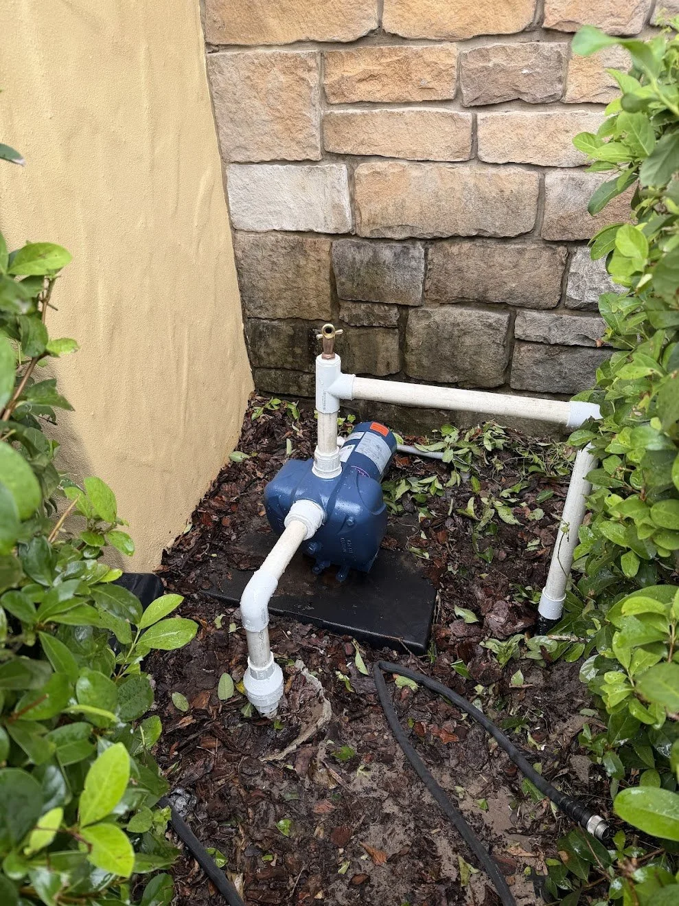 An outdoor water pump installed on the ground surrounded by green bushes and nearby brick wall.