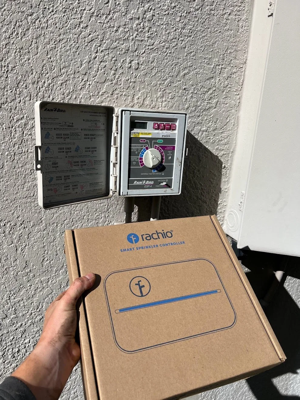A person holding a cardboard box labeled 'Rachio Smart Sprinkler Controller' stands in front of an outdoor irrigation timer mounted on a textured wall, with a white electrical panel nearby.