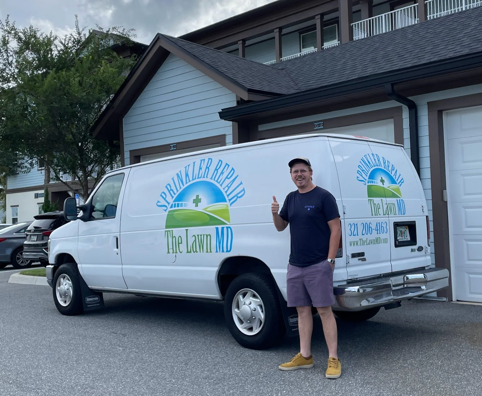 A man standing next to a white service van with the logo and contact information for Sprinkler Repair The Lawn MD. The man is smiling and giving a thumbs-up. The background shows residential buildings and parked cars.