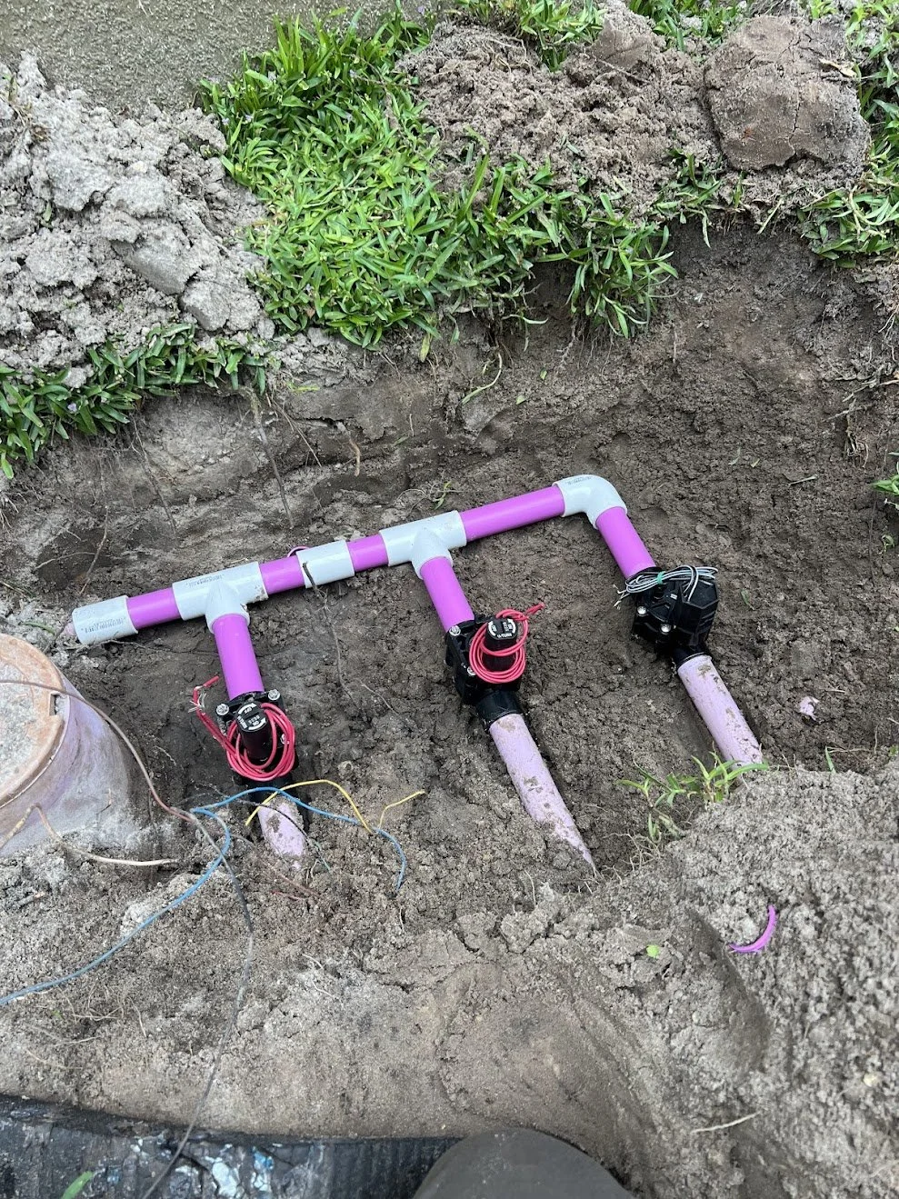 Underground irrigation system installation with purple PVC pipes, valves, and wiring, in a dug trench with soil and nearby greenery.