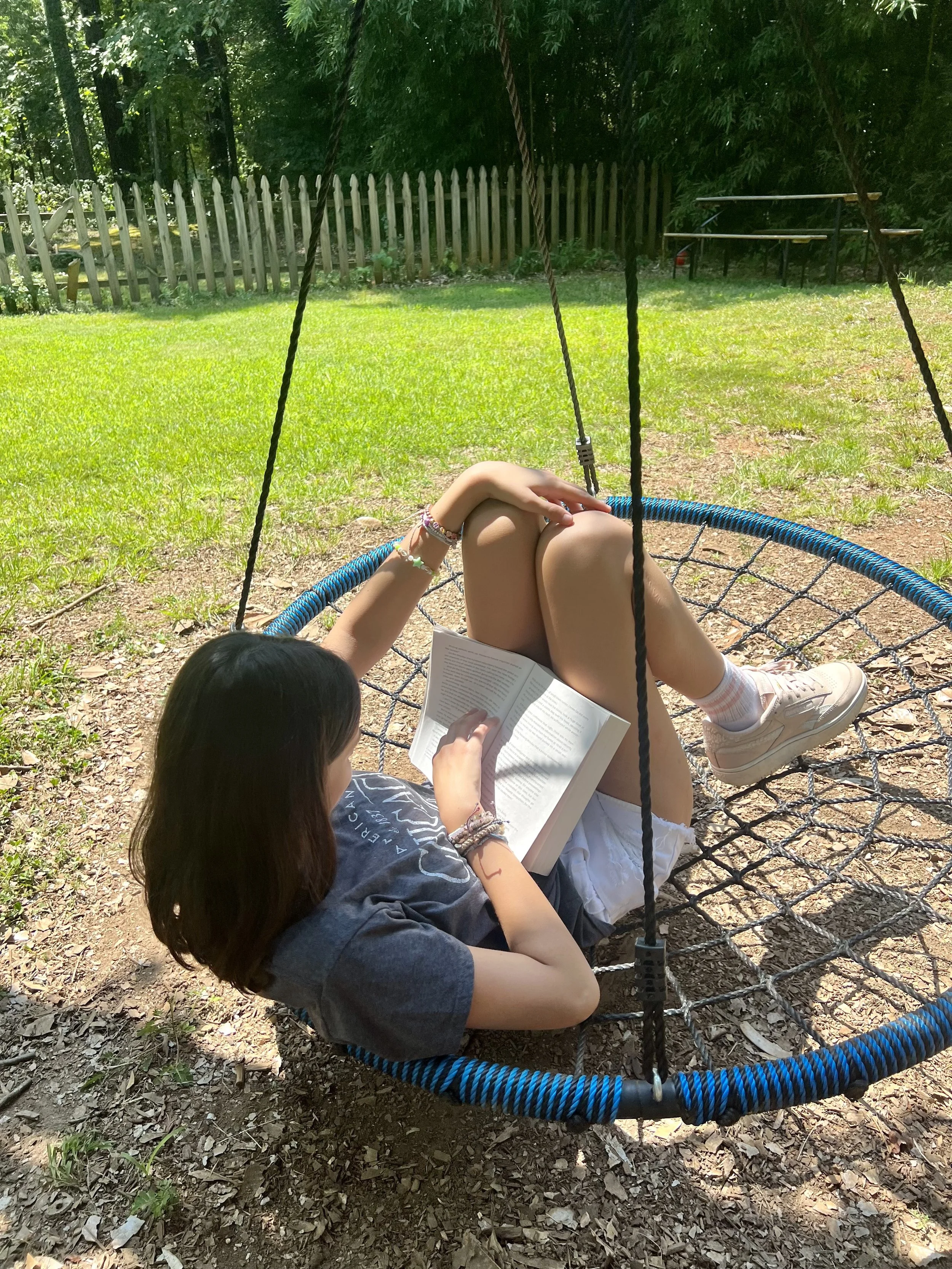 A girl relaxing on a round outdoor swing, reading a book, in a grassy backyard with trees and a wooden fence.