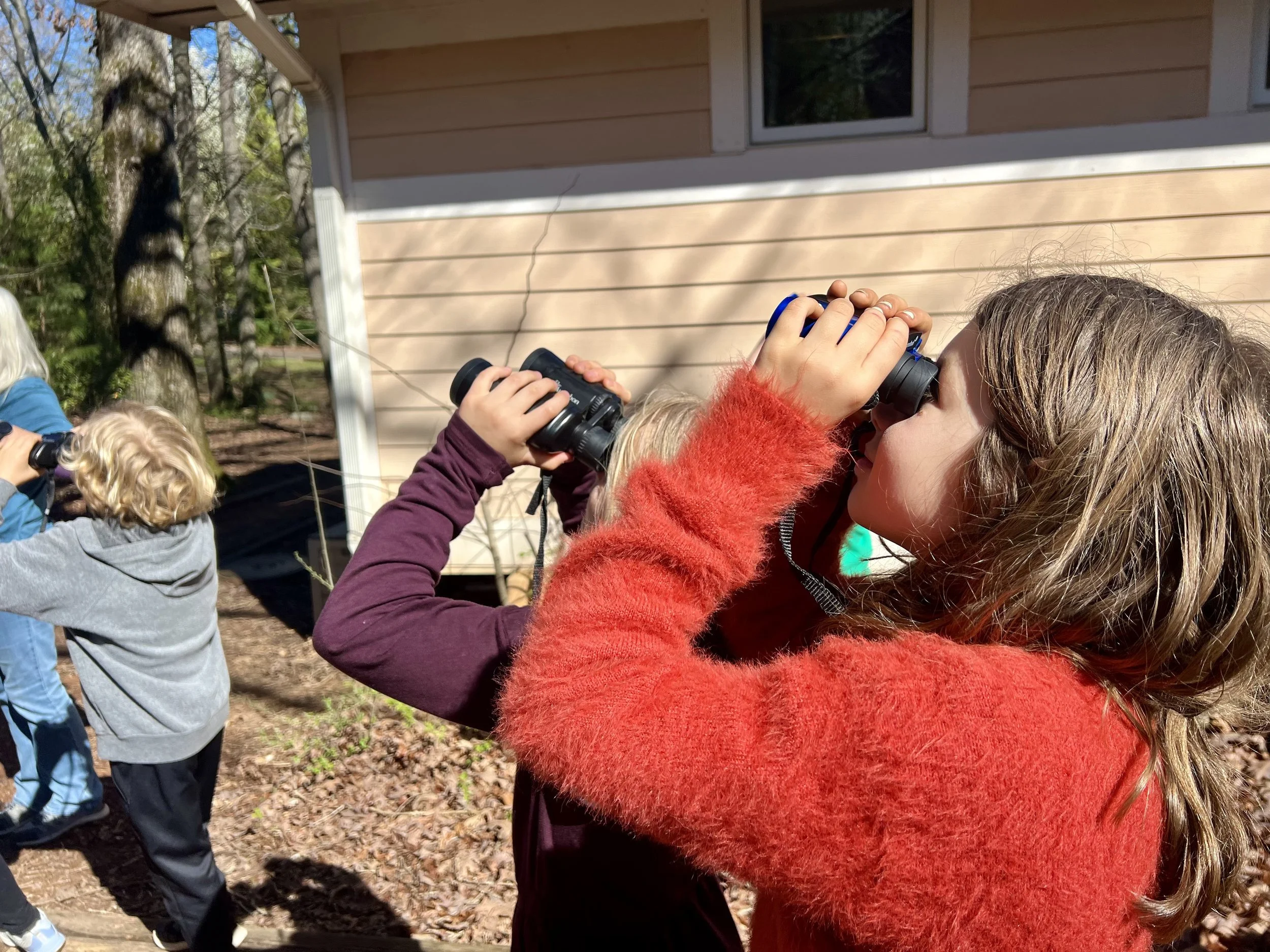 Children looking through binoculars outdoors in a wooded area.