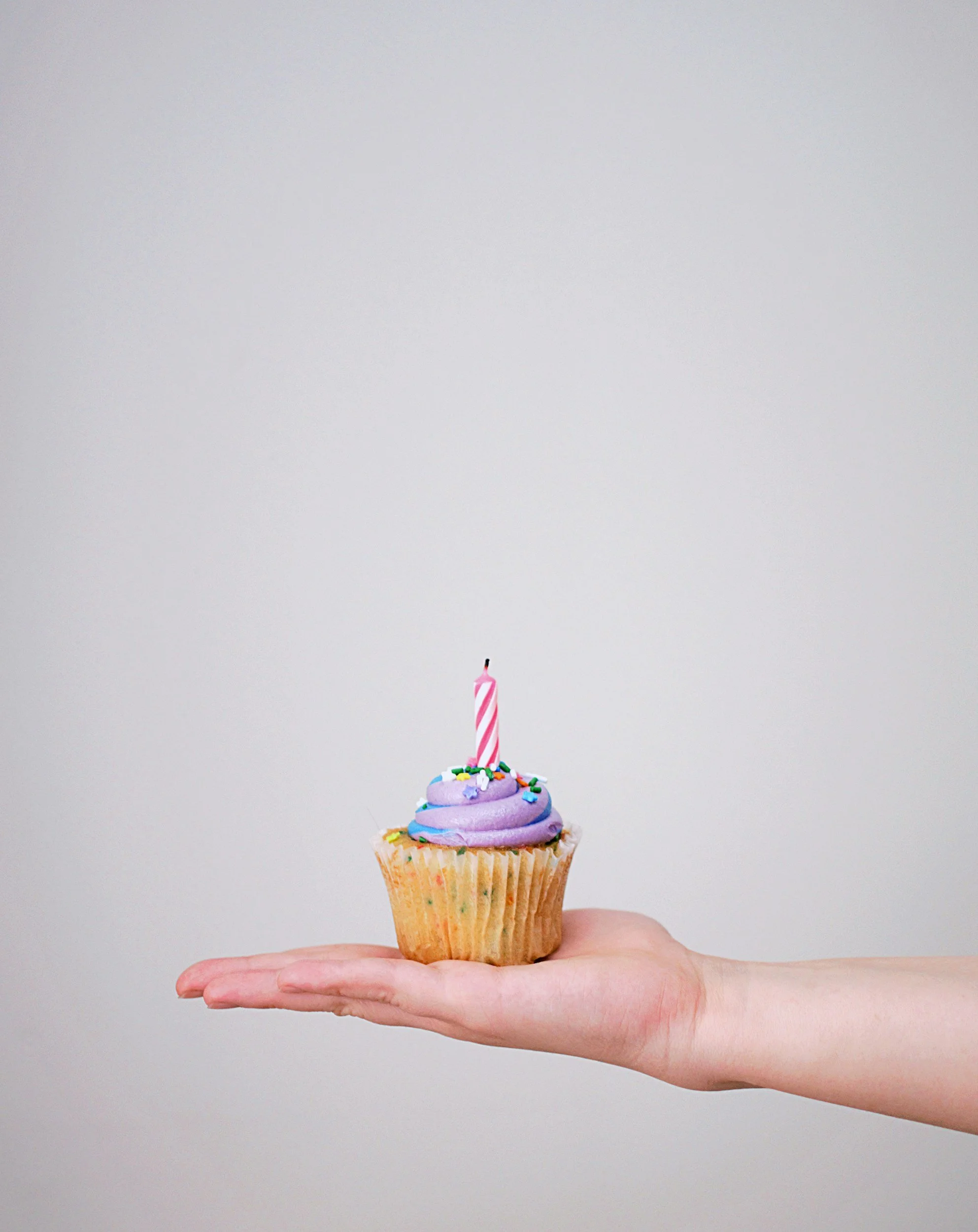 Hand holding a birthday cupcake with purple frosting, sprinkles, and a pink and white striped candle.