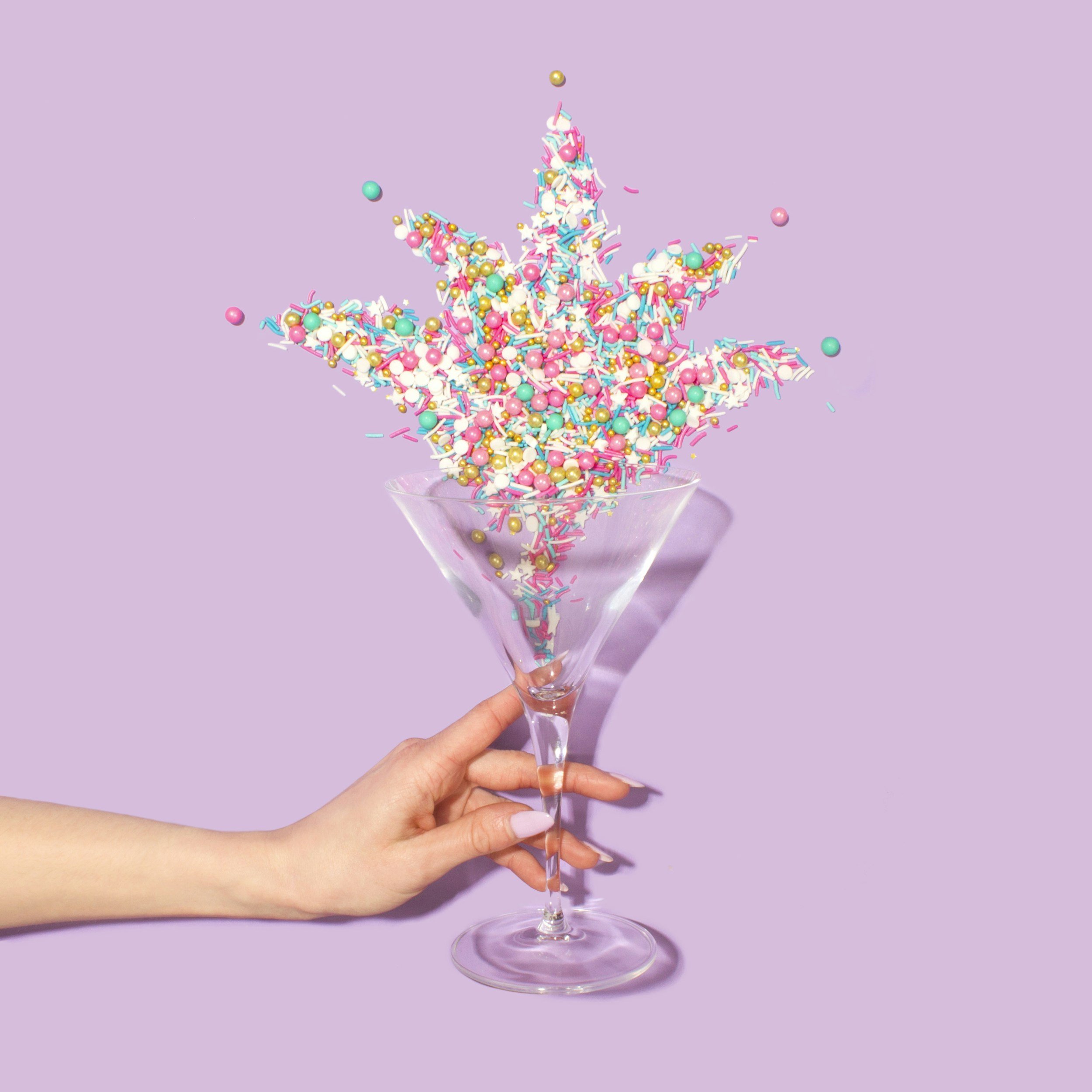 A hand holding a martini glass with colorful sprinkles and beads spilling out, forming a star shape against a pastel purple background.