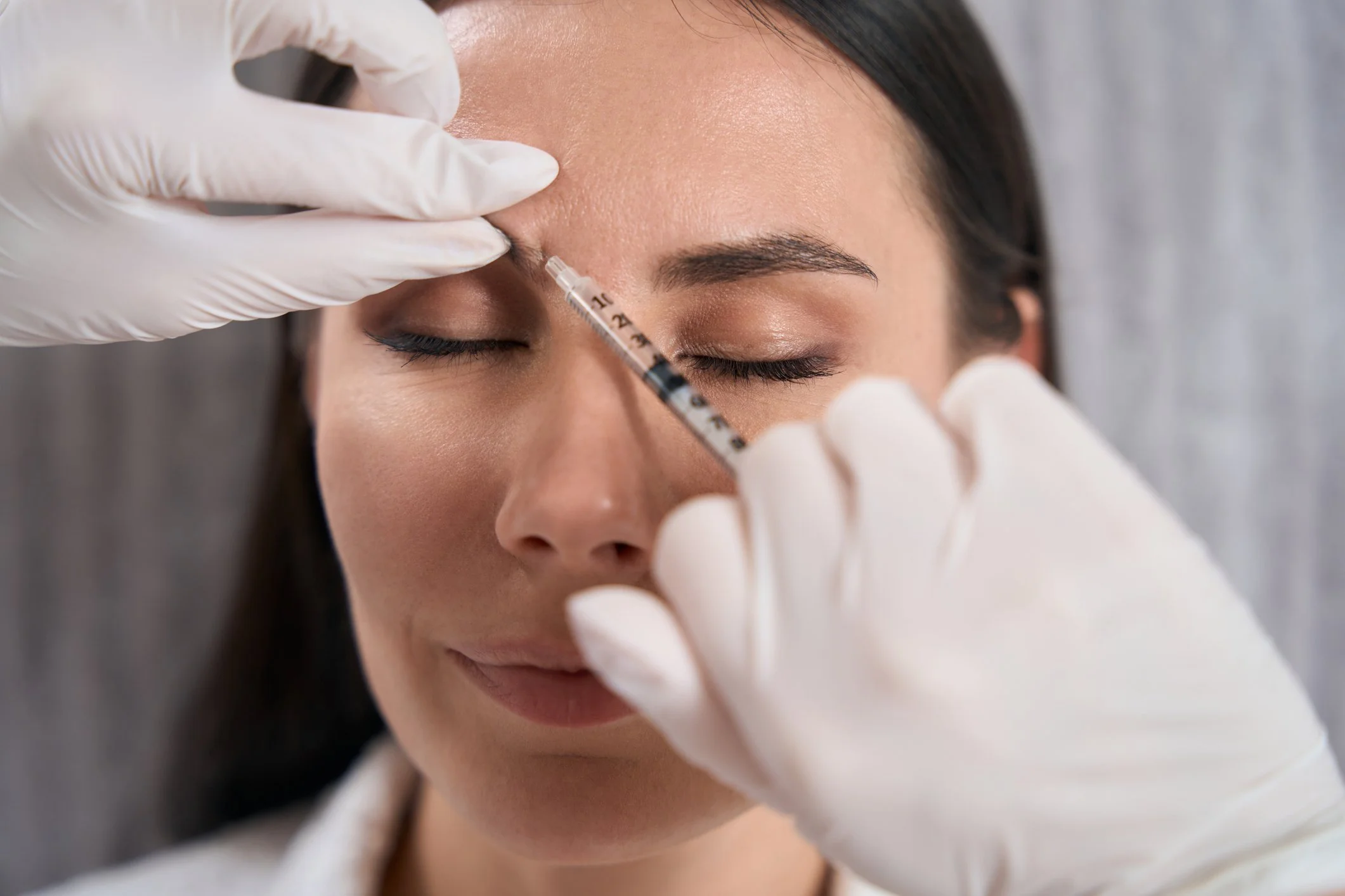 A woman with closed eyes receives a cosmetic injection in her forehead from a medical professional wearing white gloves.