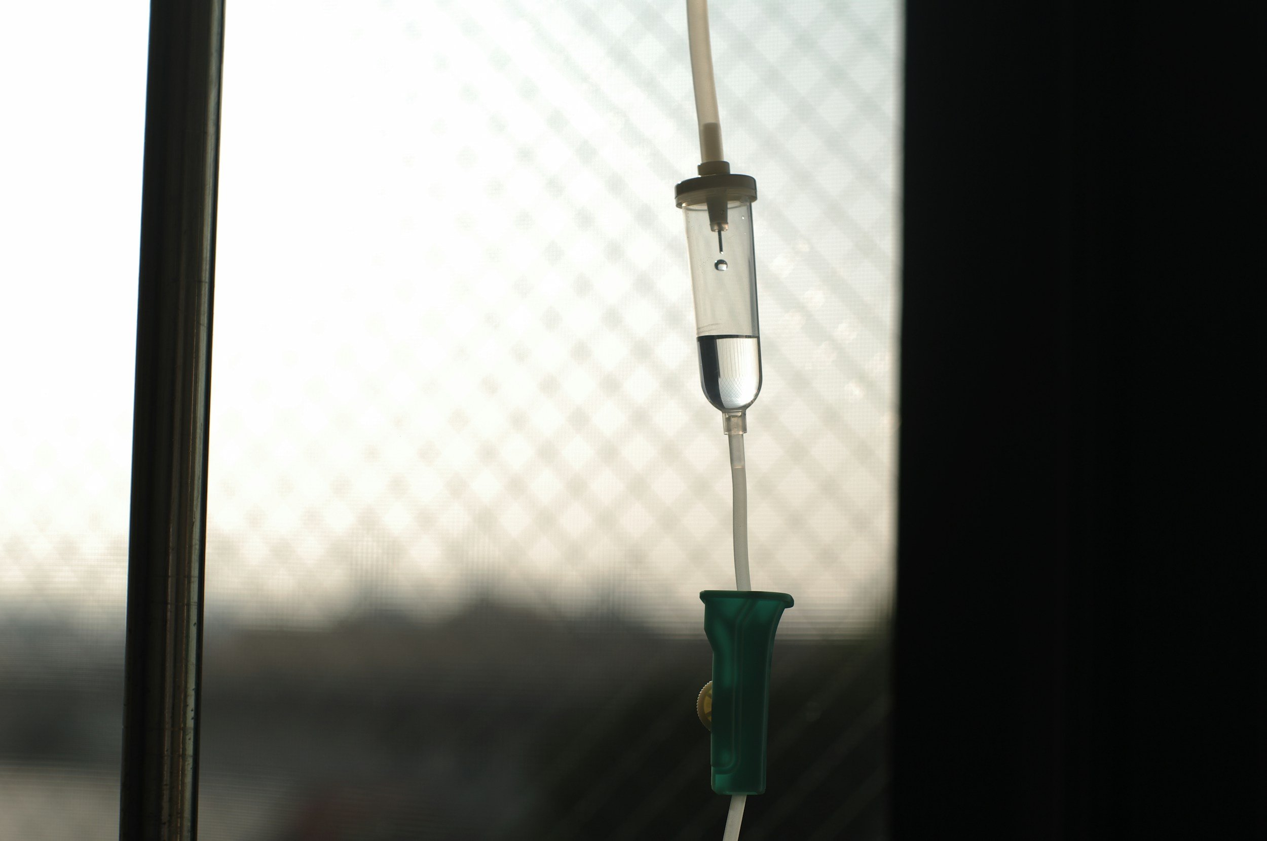 Medical IV drip hanging from a pole with a green clamp, against a blurred, patterned window background.