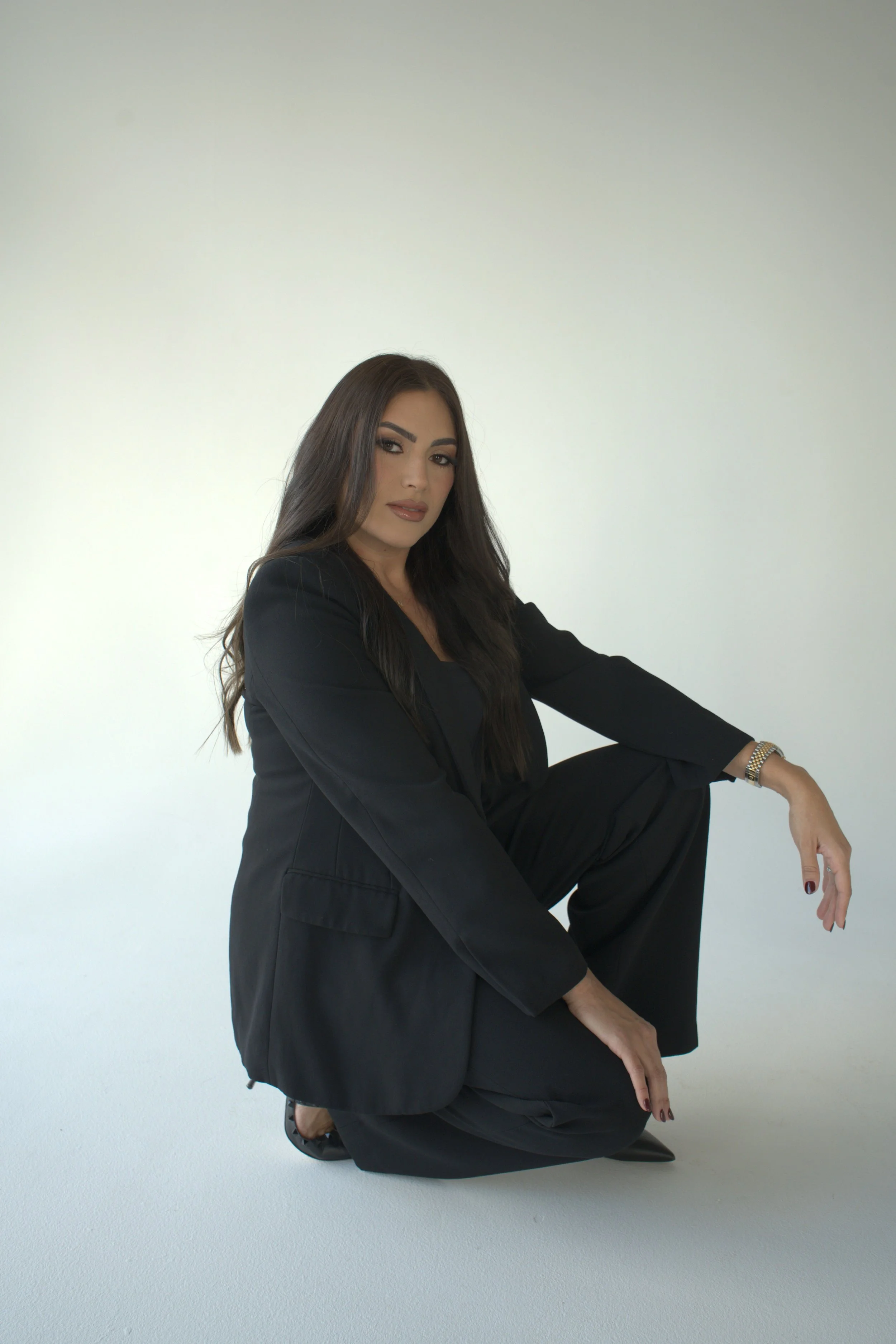 A woman in a black suit crouching on a light-colored floor against a plain light background.