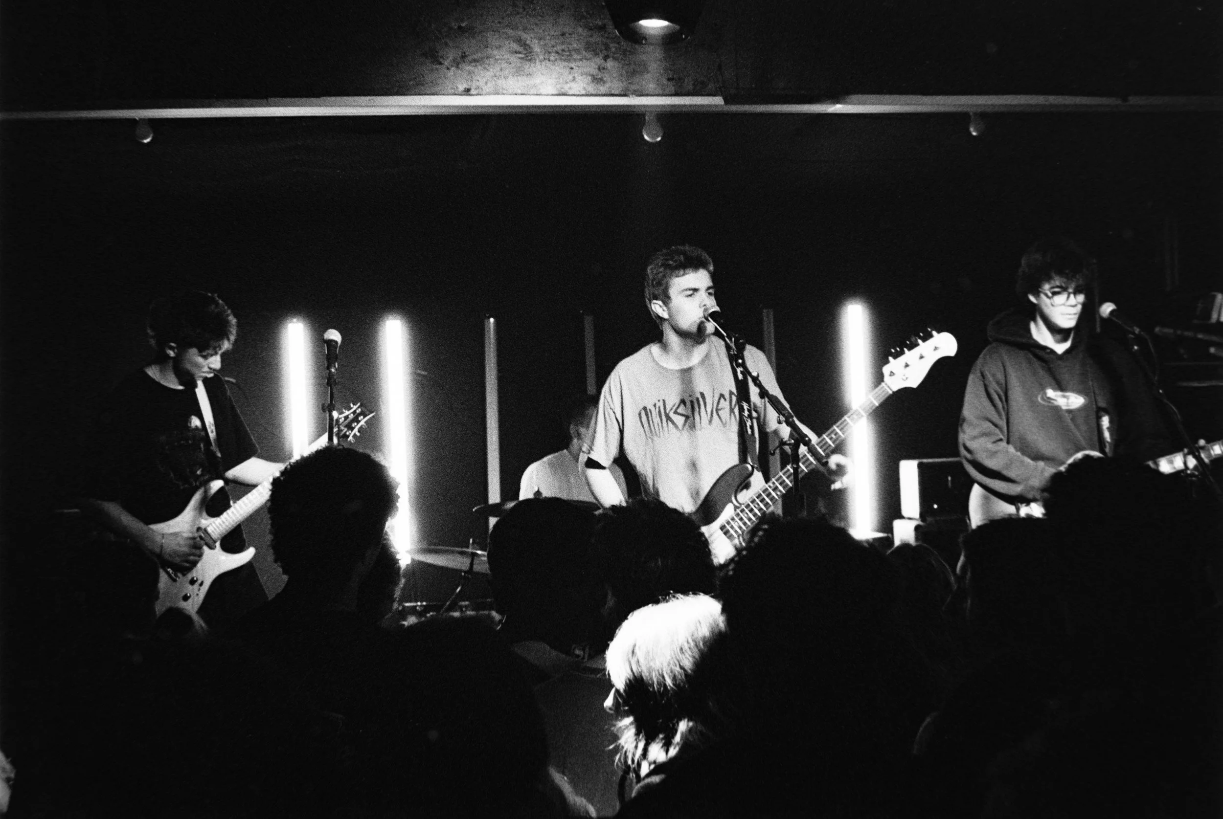 Black and white photo of a band performing on stage; three musicians with guitars and microphones, audience in foreground.