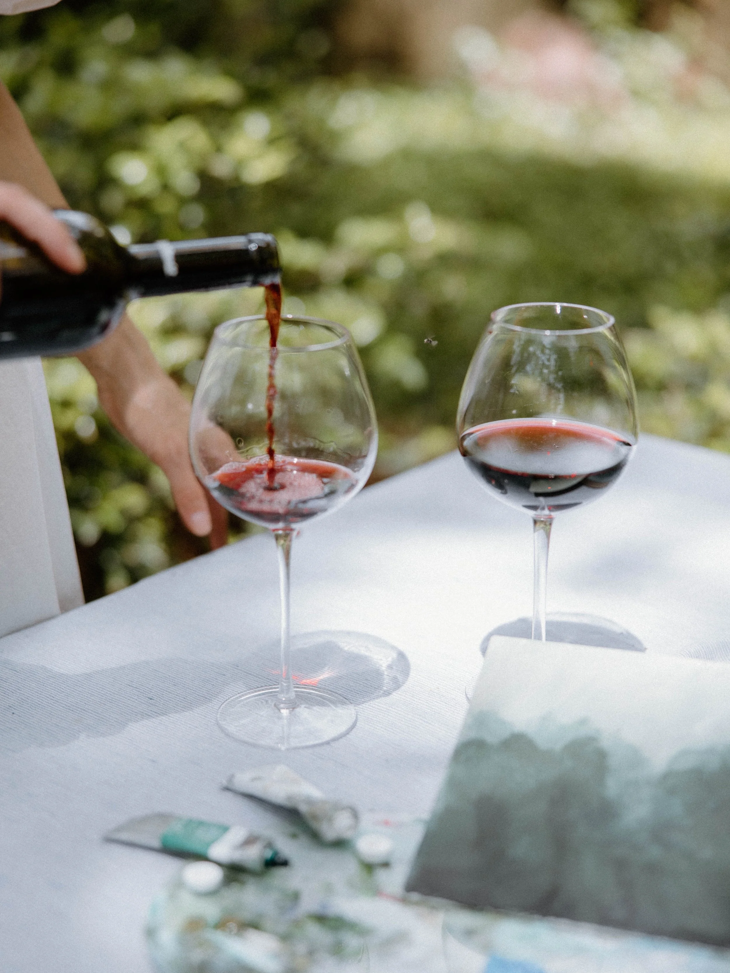 A person pouring red wine into a glass on a white table outdoors, with a second glass of wine and art supplies nearby.
