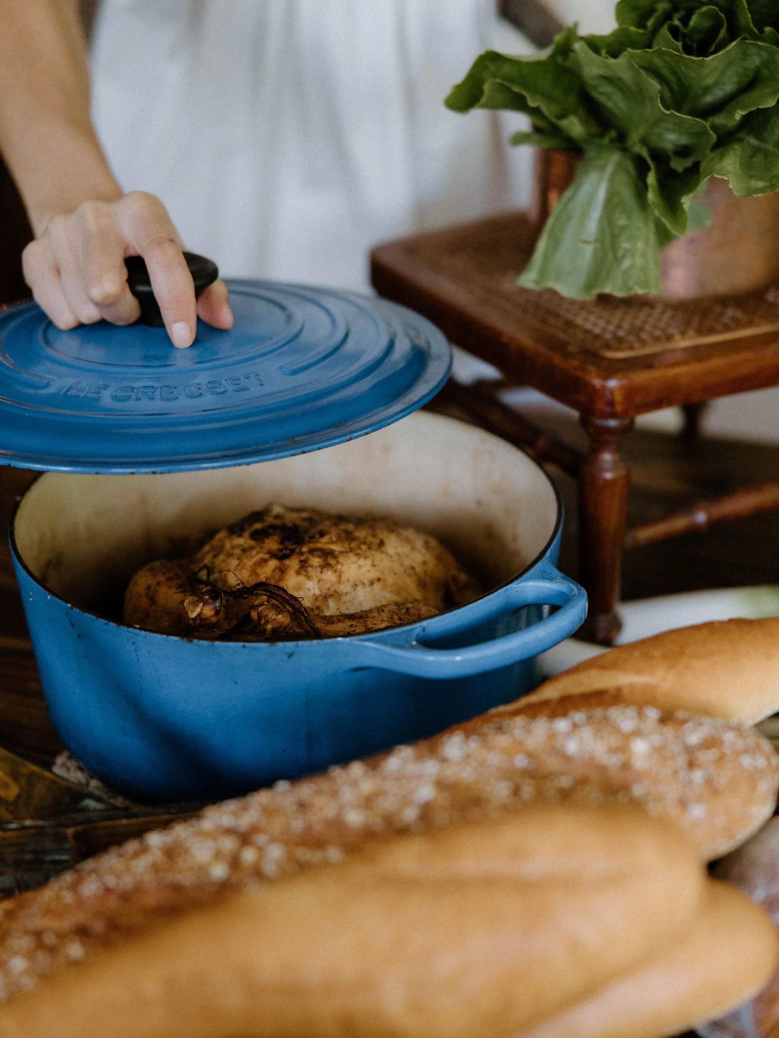 A person lifting the lid off a blue cast iron Dutch oven containing a roasted chicken, with loaves of bread in the foreground and a potted plant on a wooden table in the background.