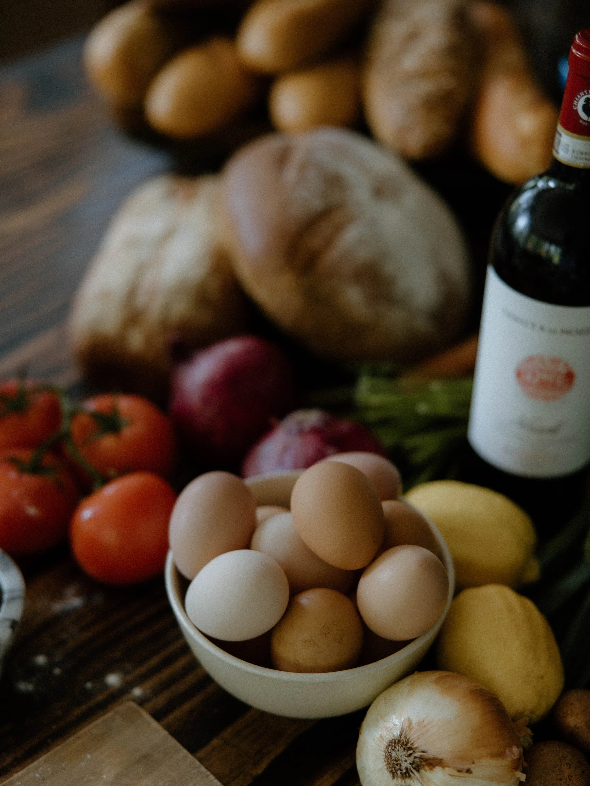 A bowl of eggs surrounded by tomatoes, lemons, onions, radishes, and potatoes on a wooden table.