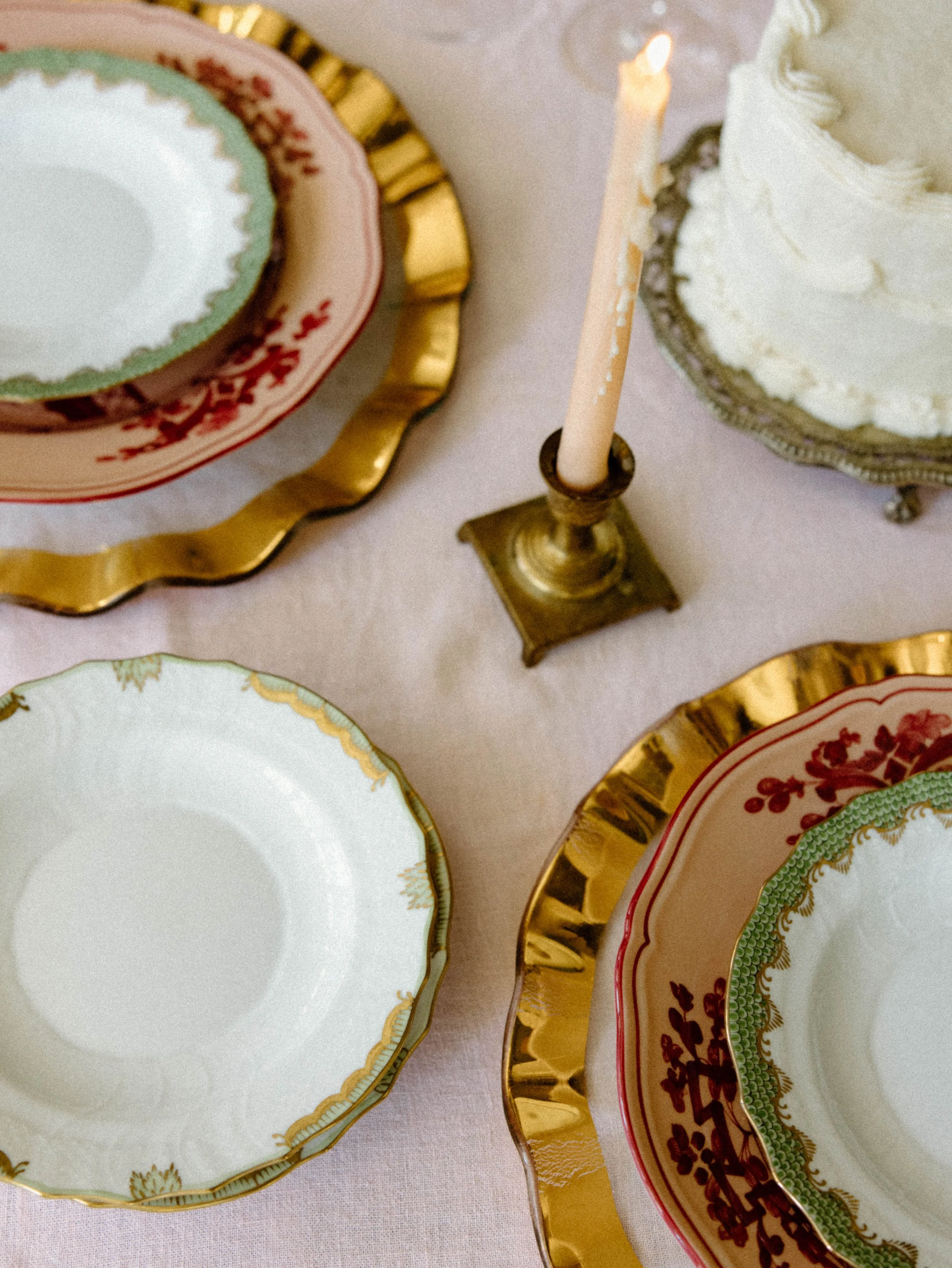 Elegant table setting with decorative plates featuring gold rims and floral patterns, a white layered cake on a silver stand, and a lit pink birthday candle on a brass holder.