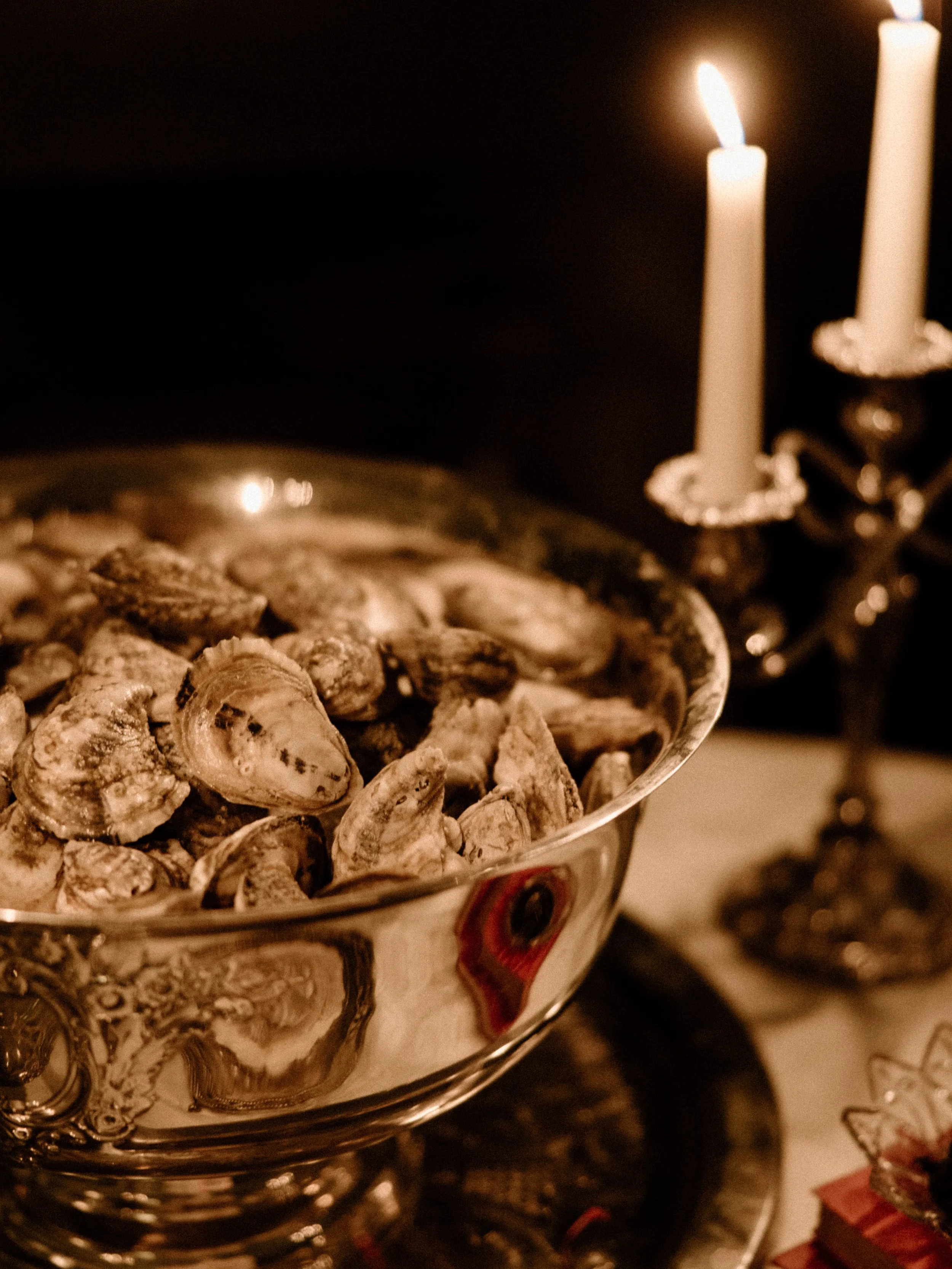 A silver bowl filled with oysters on a table illuminated by two lit candles in the background.