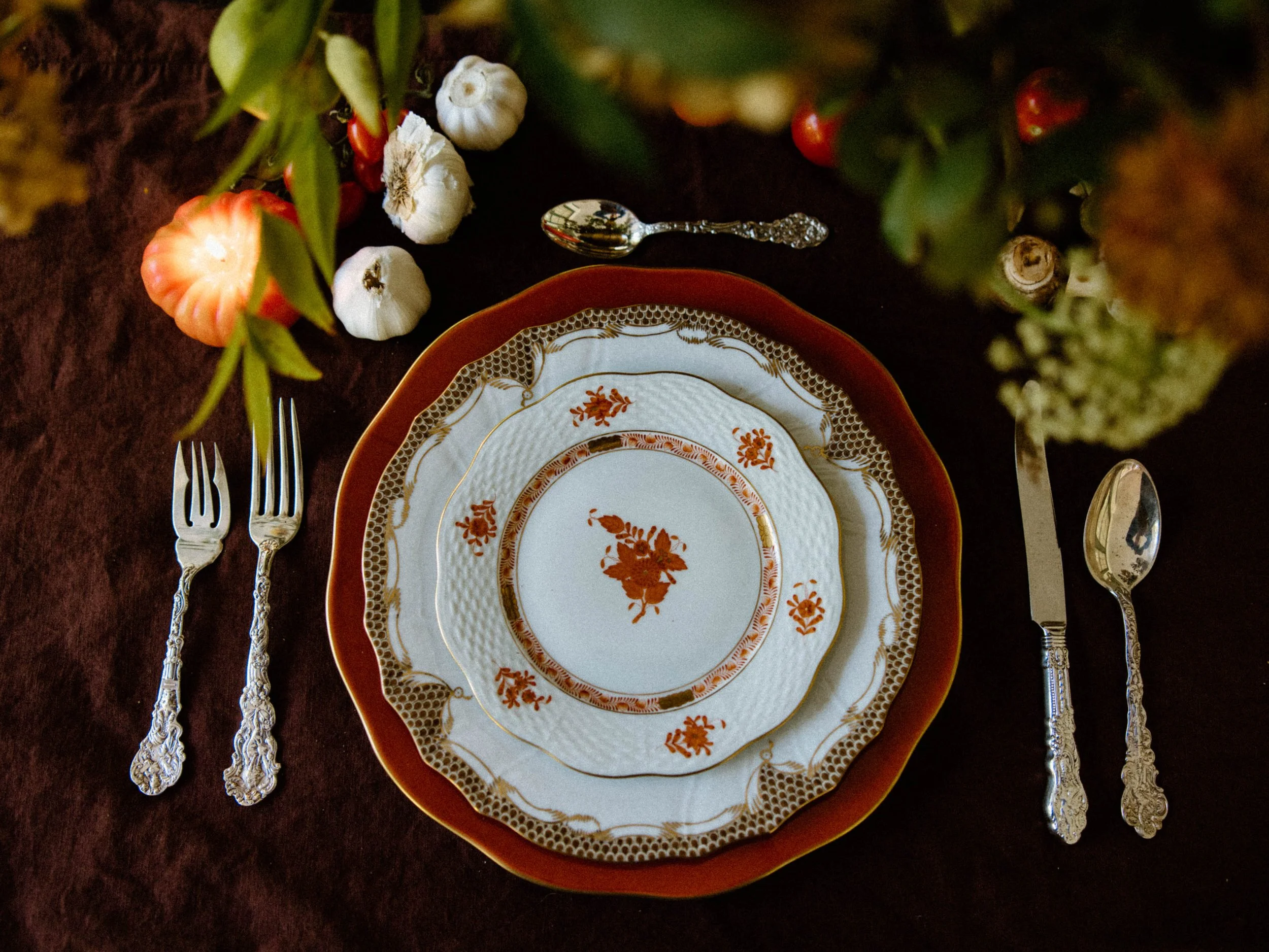 Elegant table setting with vintage china plates, silverware, small decorative gourds, garlic bulbs, and green foliage on a dark tablecloth.