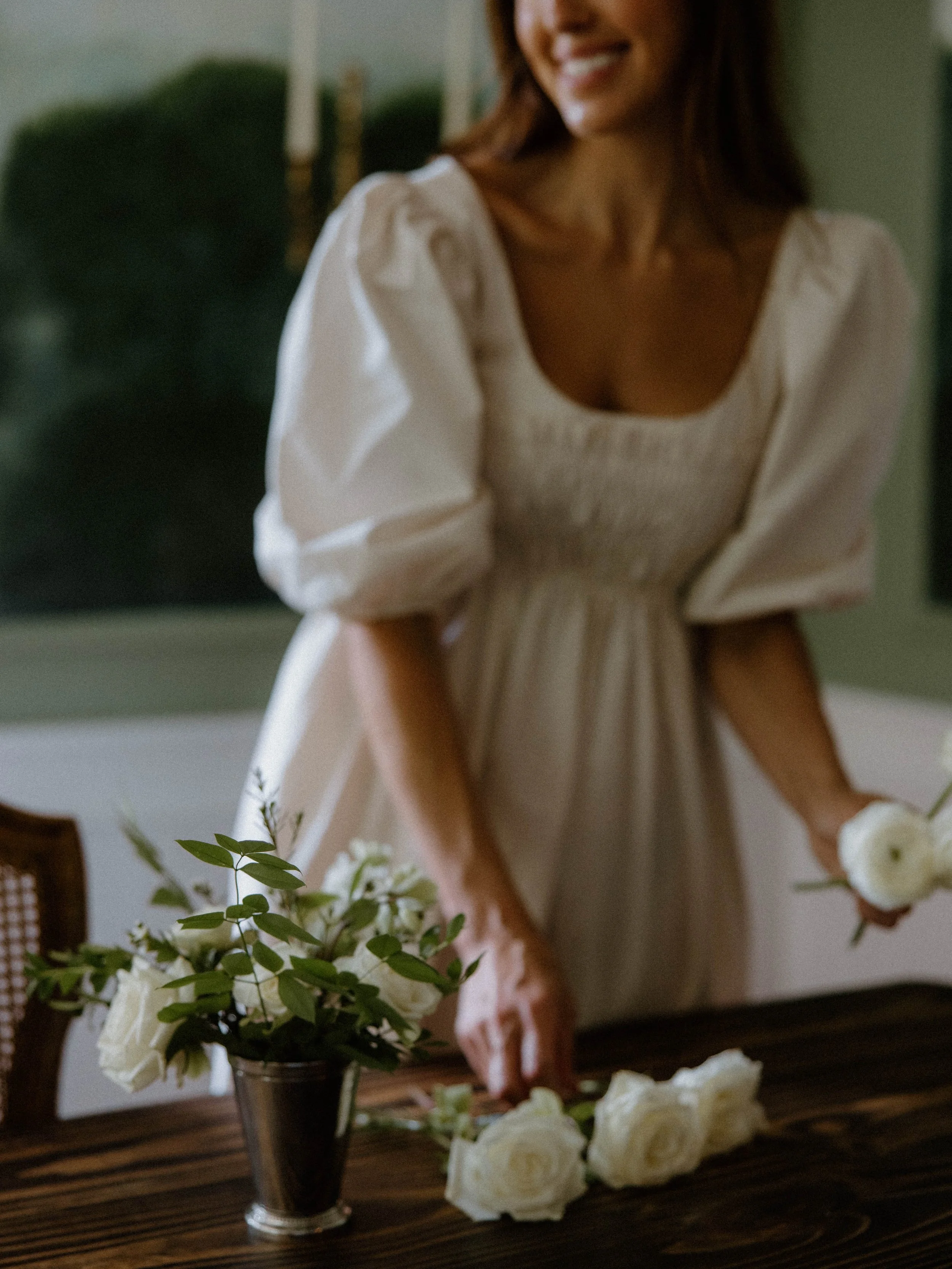 A woman is arranging white roses in a vase on a wooden table, smiling and wearing a cream-colored dress with puffy sleeves.