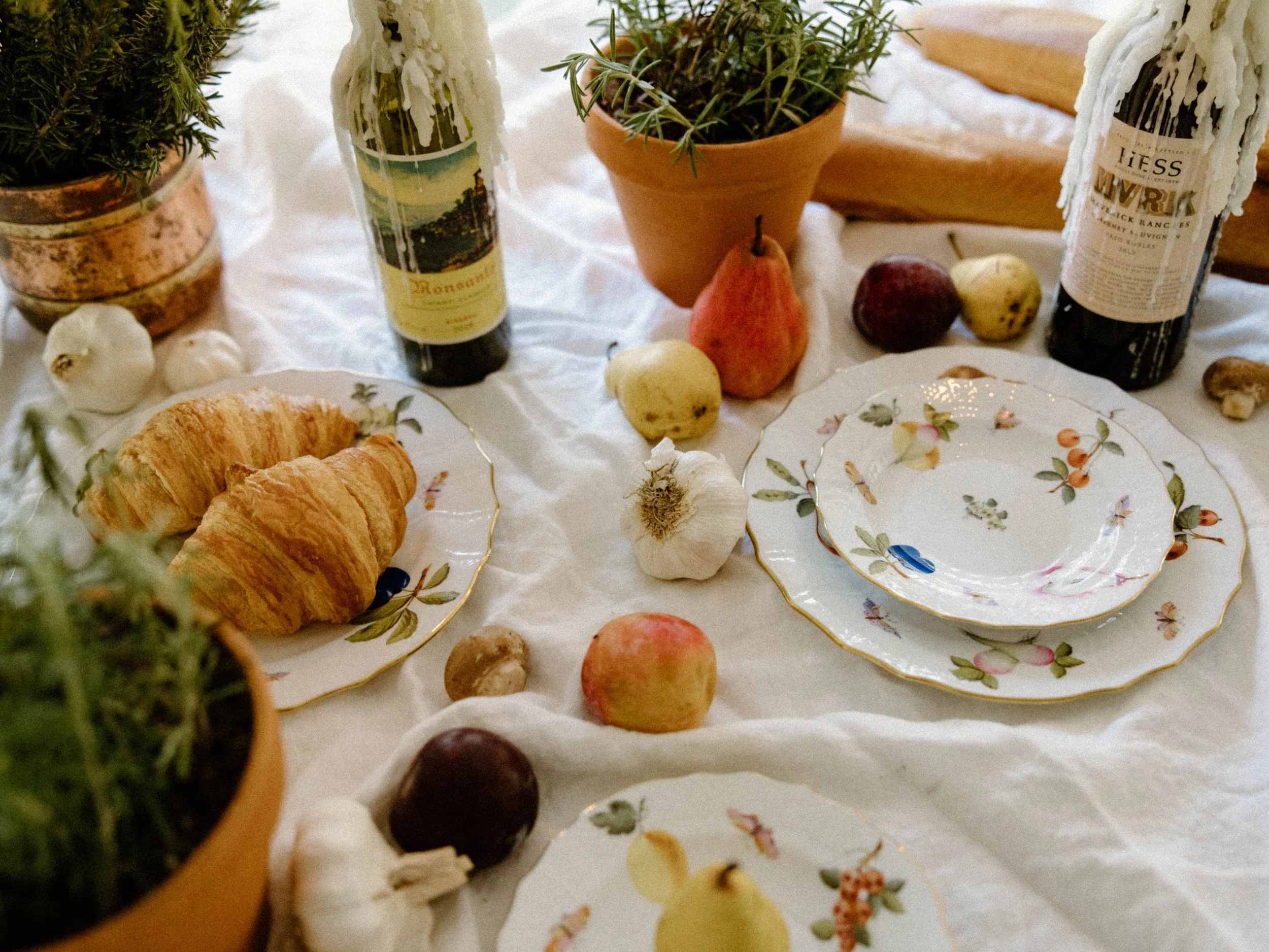 A table decorated with flowers, fresh fruits, garlic, bread, and bottles of wine, set with floral patterned plates, on a white tablecloth.