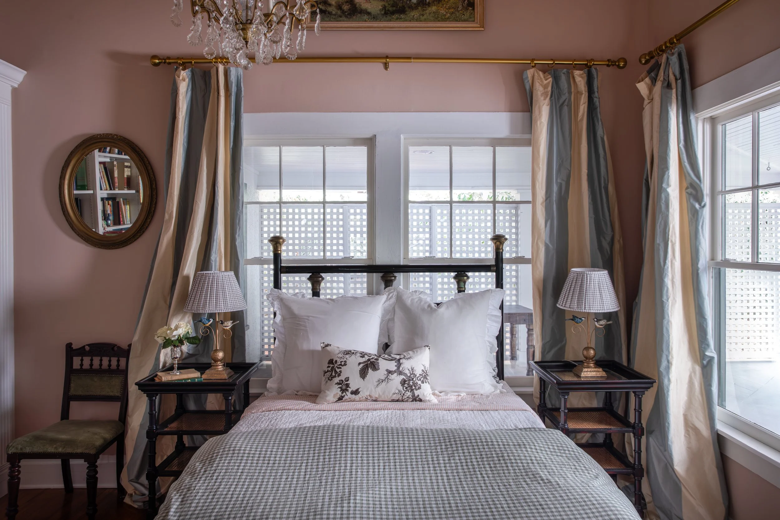 A cozy bedroom with a bed featuring white pillows and a decorative pillow, flanked by two black nightstands with lamps, surrounded by large windows with curtains, a chandelier hanging from the ceiling, and a bookshelf with books reflected in a round