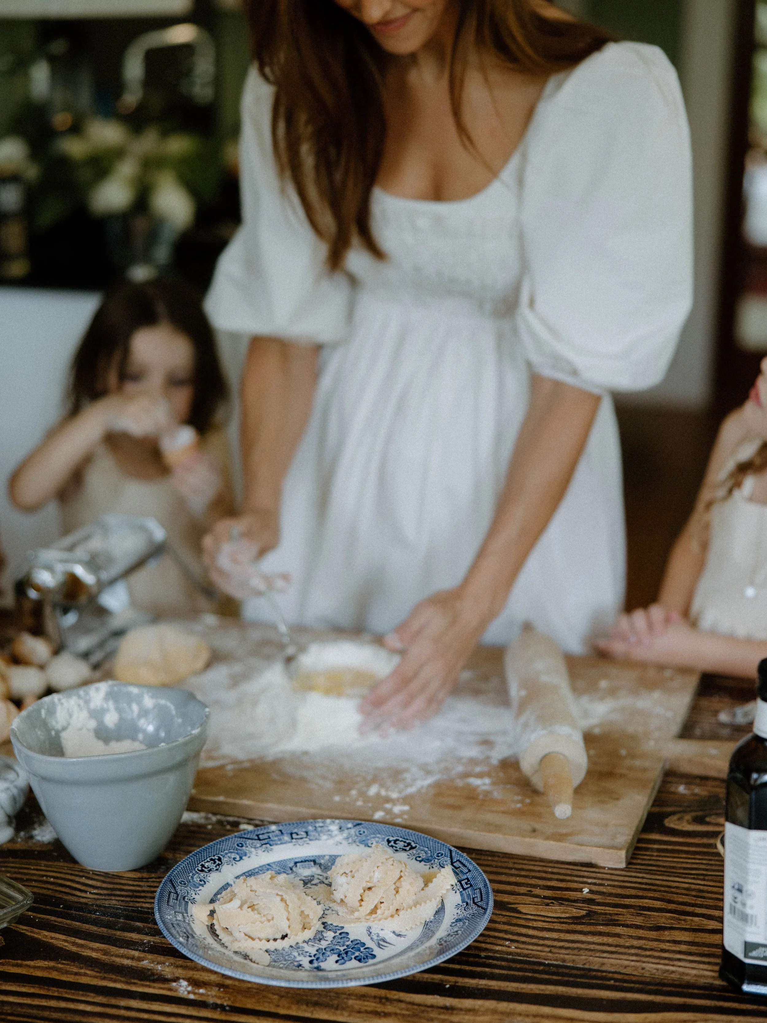 Woman and two children making homemade pasta together in a kitchen, with pasta dough, rolling pin, and ingredients on the wooden table.