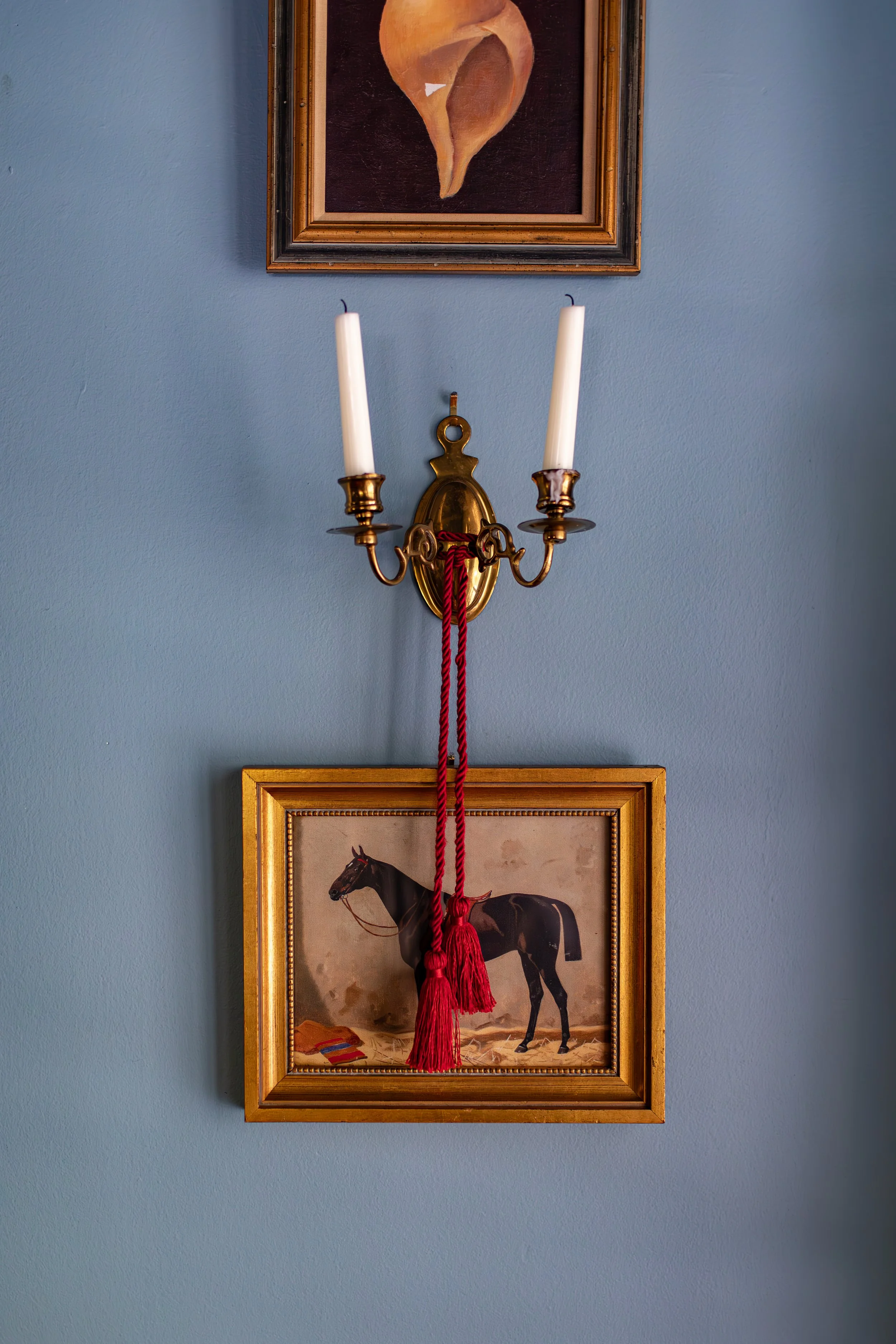 A wall-mounted brass candle holder with two unlit white candles, a framed painting of a seashell above it, and a framed painting of a black horse below, with a red tassel hanging from the candle holder.