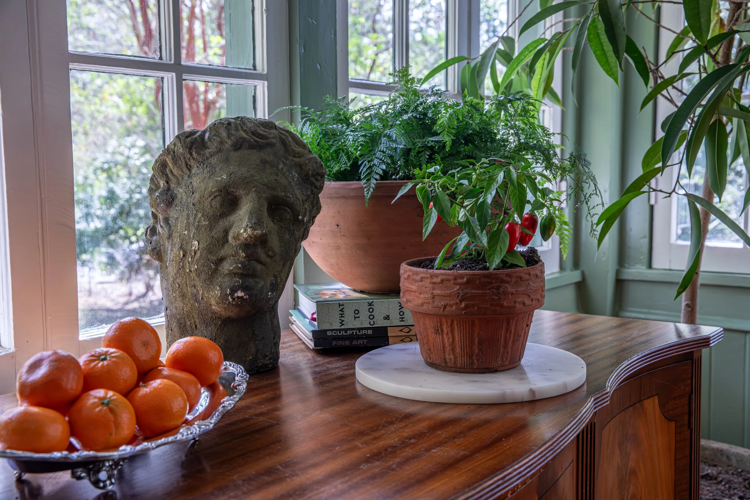Indoor wooden table with a sculpted stone head, a bowl of oranges, a stack of books, and potted houseplants near a window.