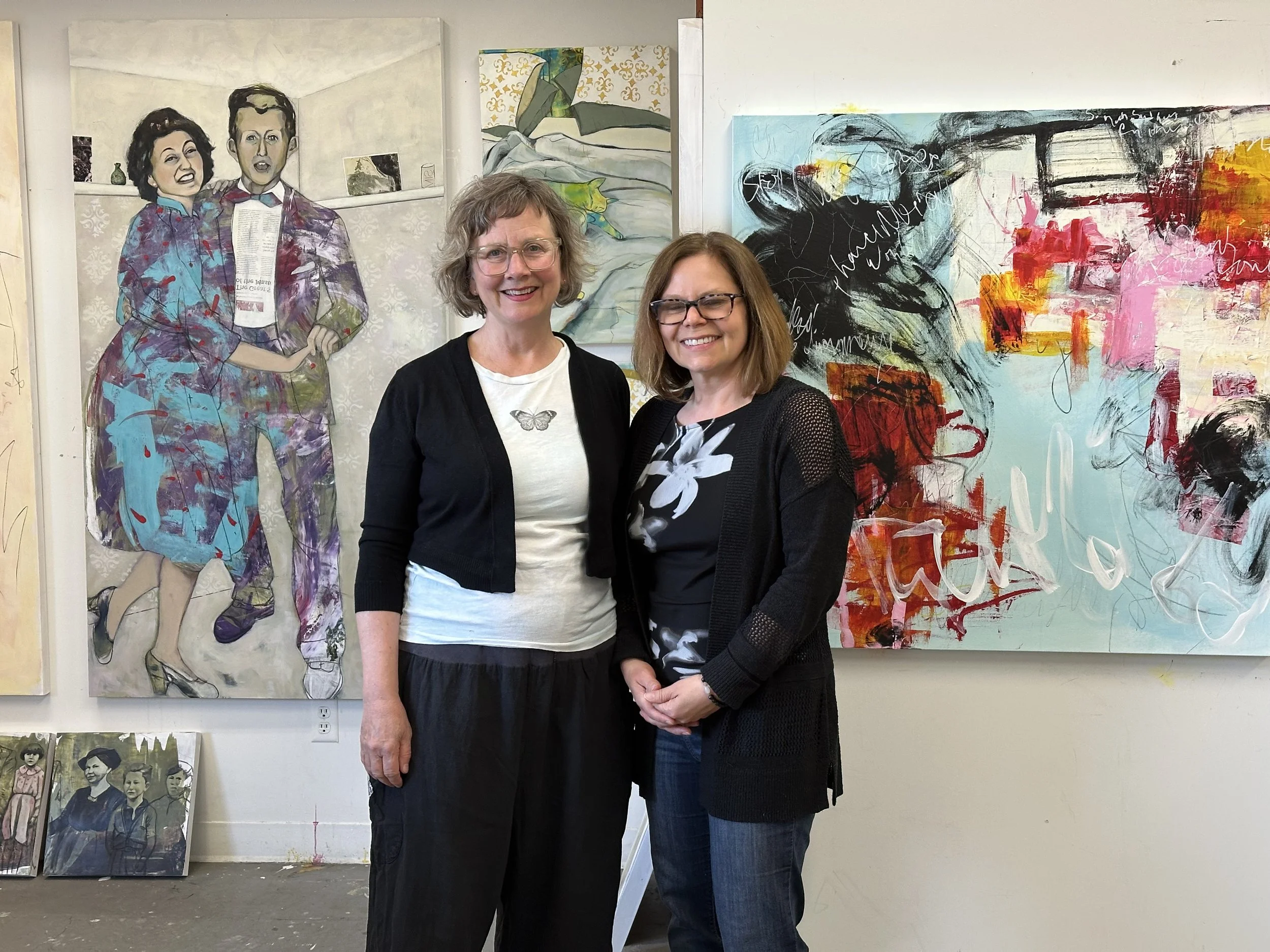 Two women standing in front of colorful abstract artwork in an art gallery. One woman has short curly gray hair, glasses, and is wearing a black cardigan over a white shirt. The other woman has shoulder-length straight brown hair, glasses, and is wea