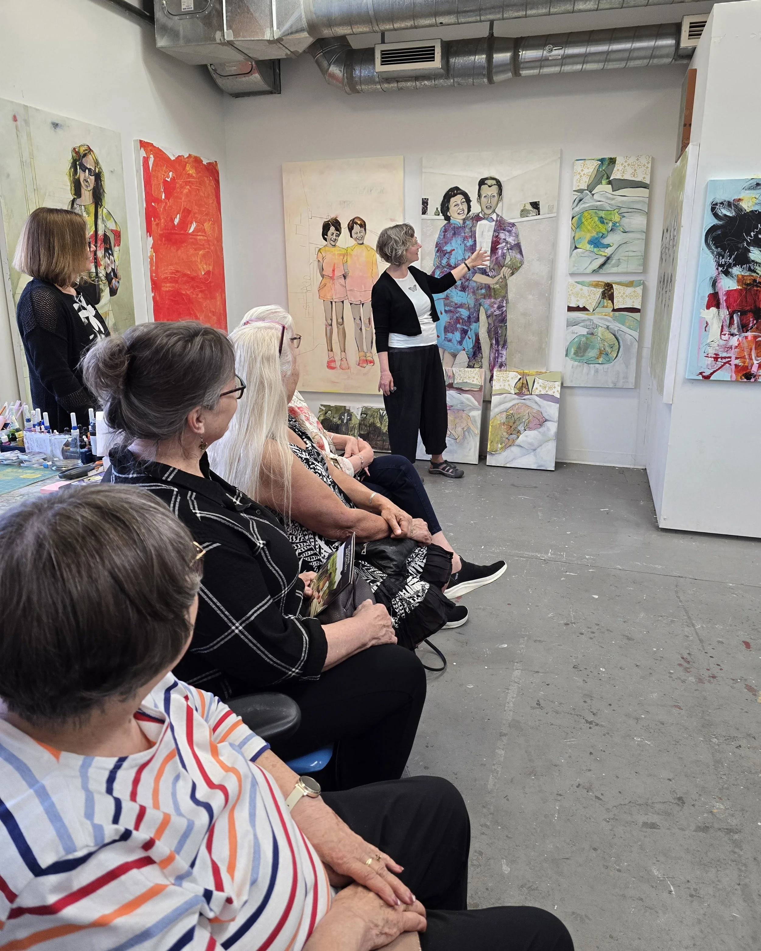 Art instructor presenting a lecture on paintings to an audience in an art gallery or studio.