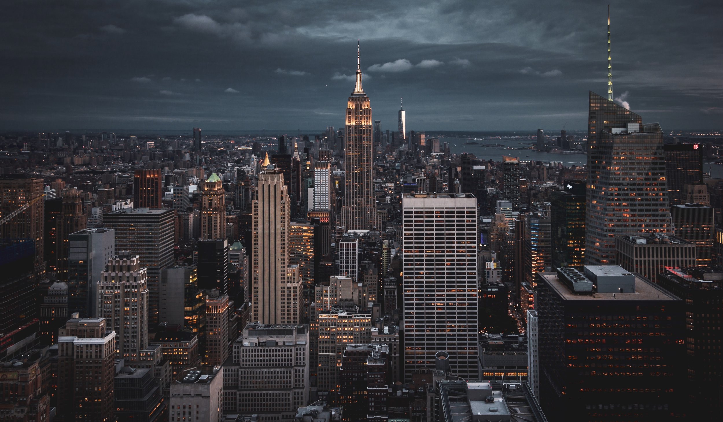 Aerial view of New York City skyline at dusk with iconic skyscrapers illuminated, including the Empire State Building, under a cloudy sky.