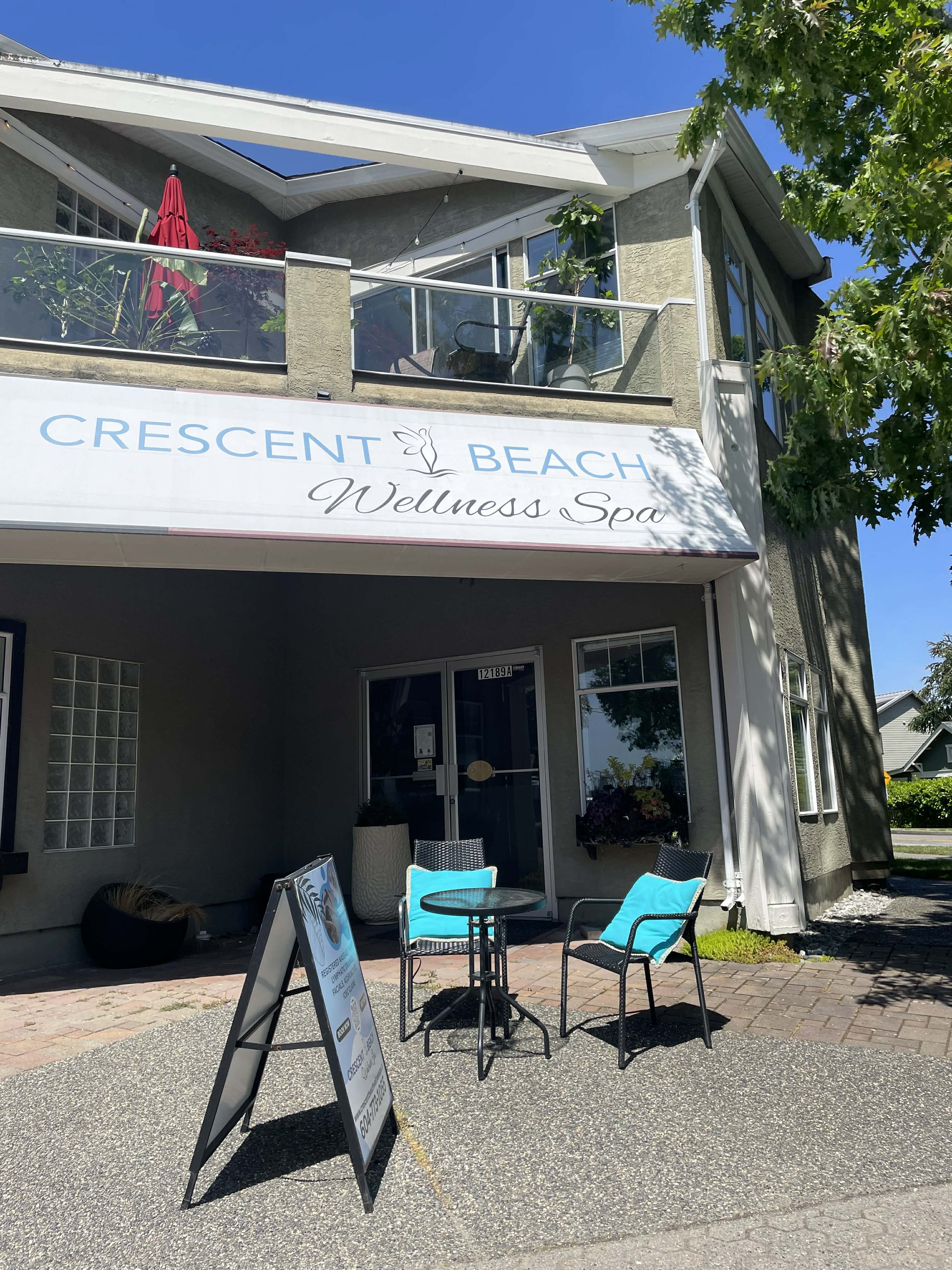 Outside view of Crescent Beach Wellness Spa with patio furniture, a signboard, and a building with balcony and large windows, under a clear blue sky with green trees.