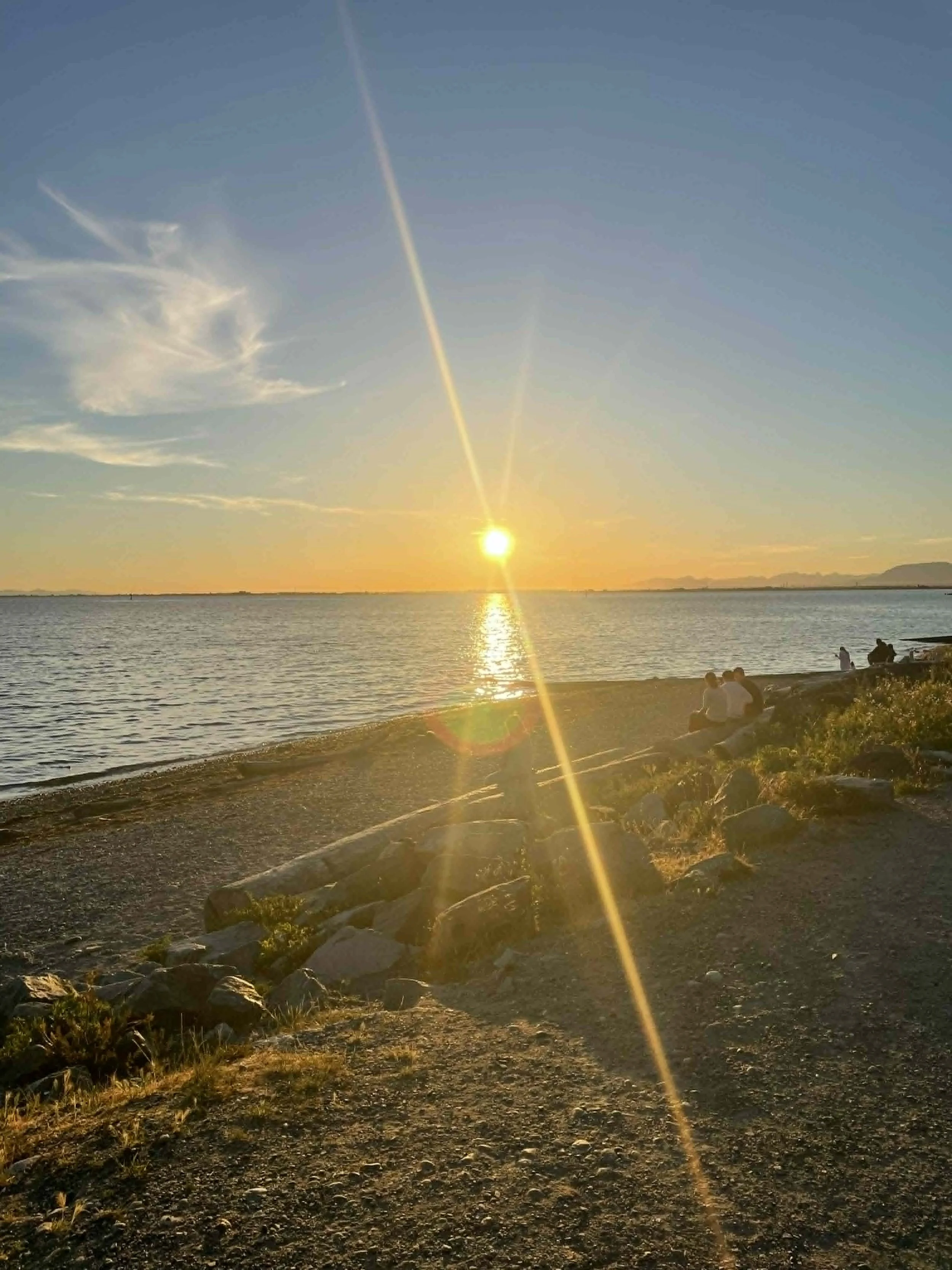 Sunset over a calm body of water with a few people sitting on rocks near the shore, and a partly cloudy sky with a visible contrail.