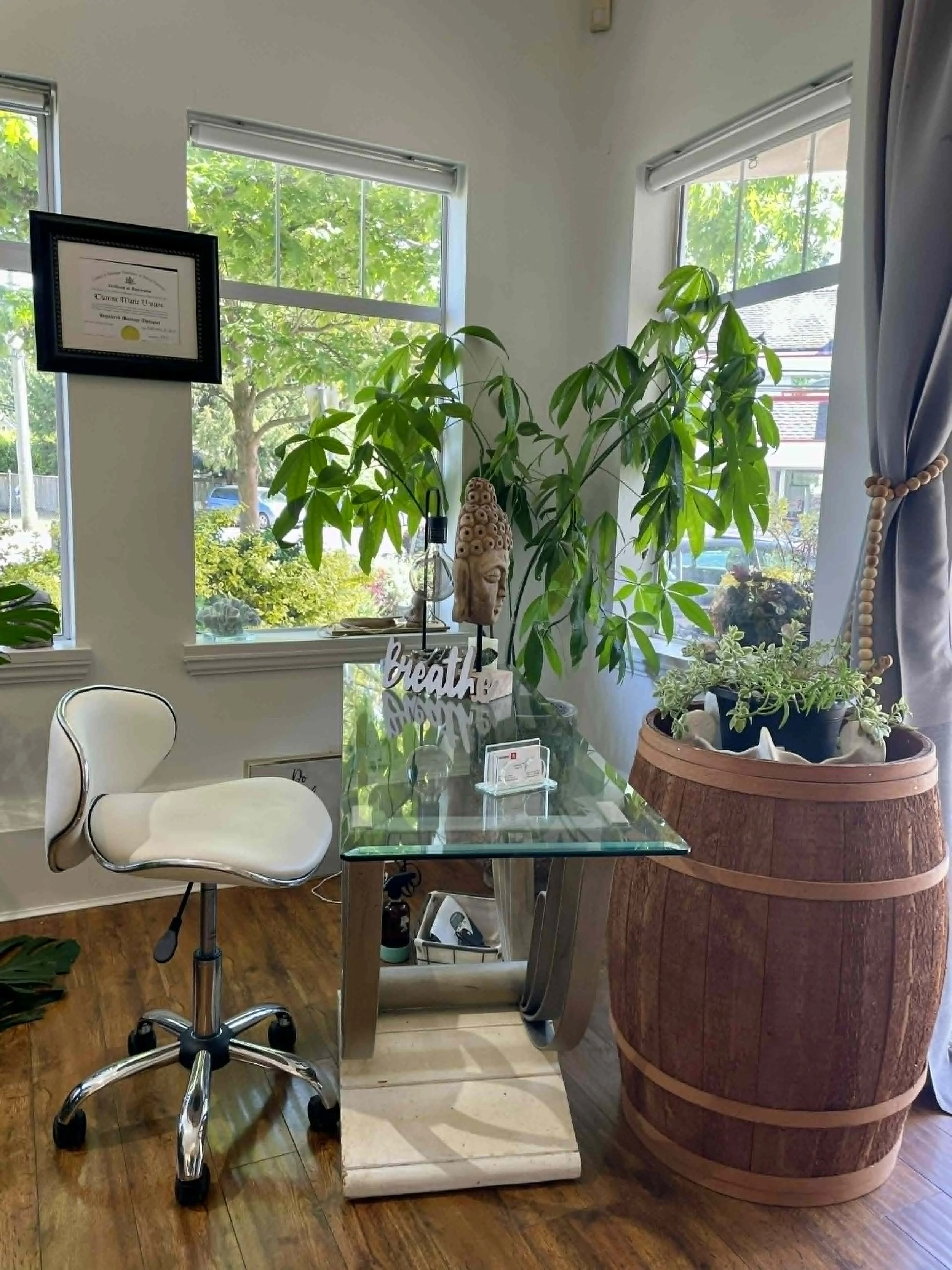 A dining room corner with a glass table, a white swivel chair, a potted plant, a Buddha statue, and windows with greenery outside.