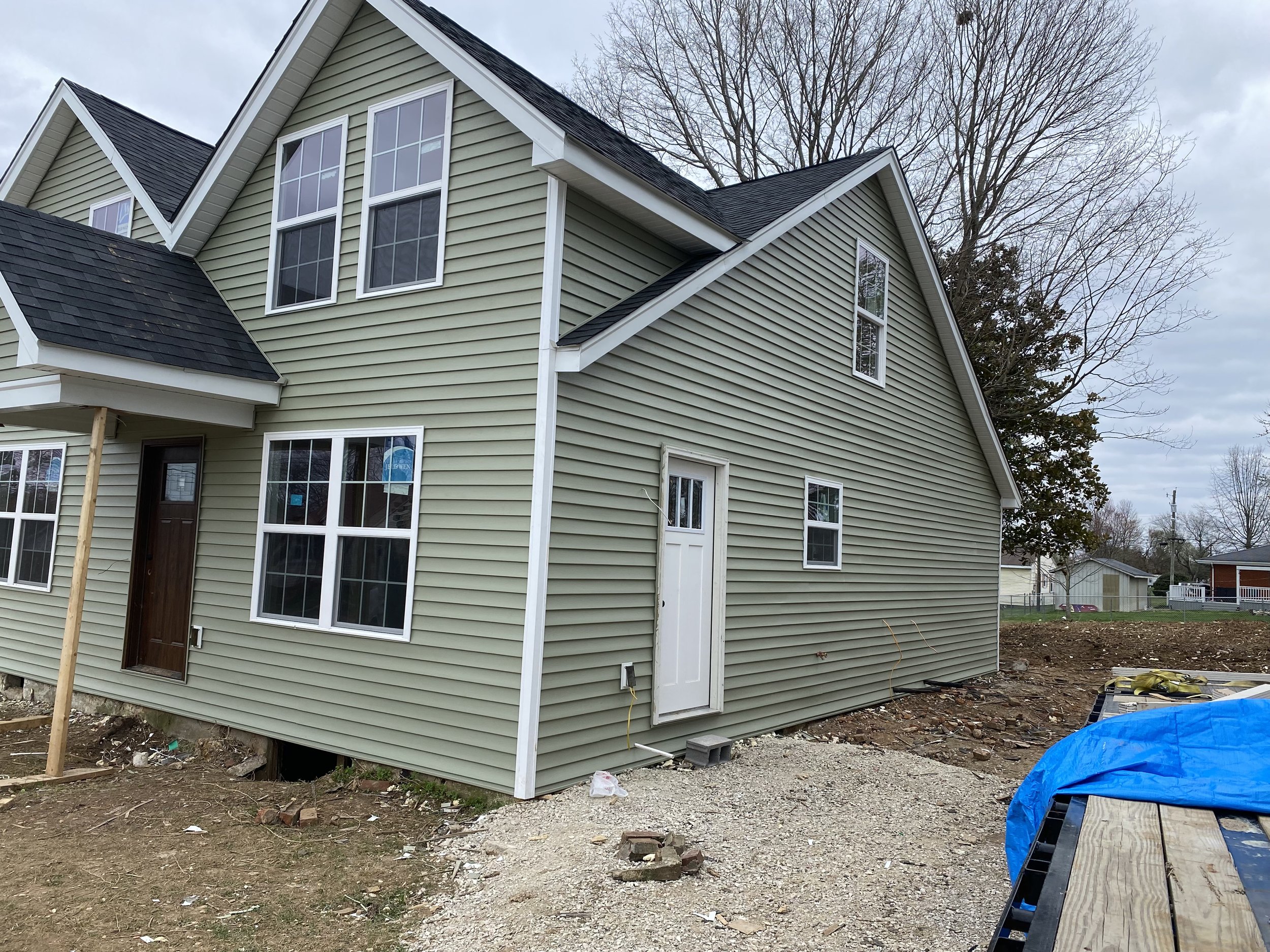 A newly constructed two-story house with green vinyl siding and asphalt shingle roof. The house features multiple windows and a wooden front door. A small side door is visible. The yard is unfinished with gravel and construction materials.