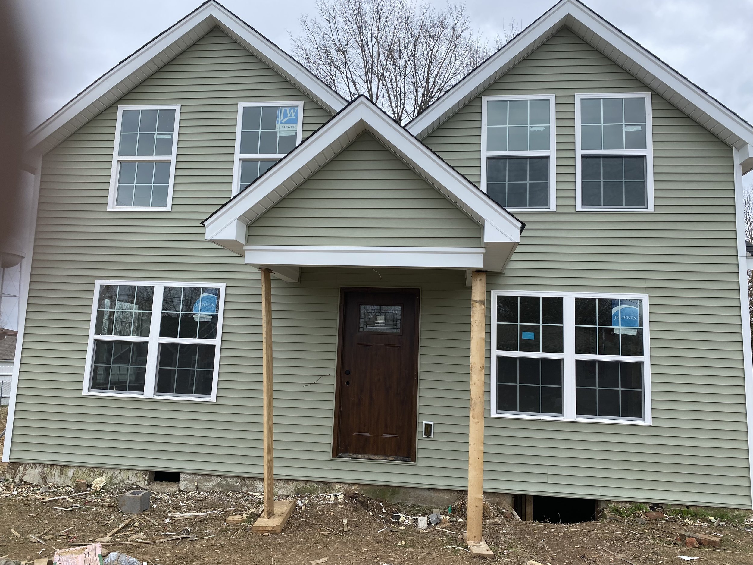 Two-story house under construction with green siding, multiple large windows, and a brown front door. The house has a gabled roof and a small porch supported by wooden posts. The ground around the house is bare with some construction debris.