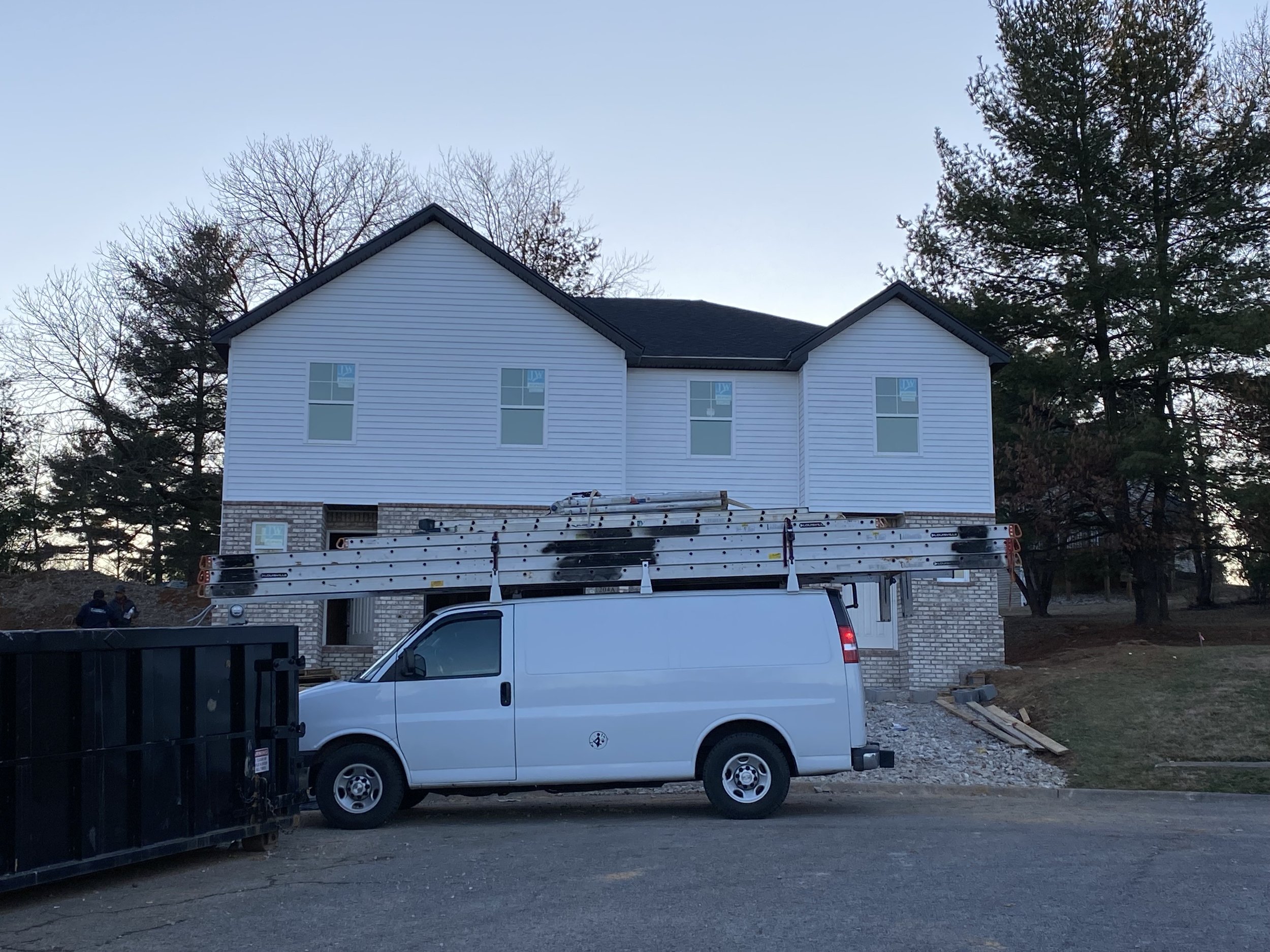 House under construction with white siding, ladders on roof, white van parked in front, surrounded by trees and a dumpster.