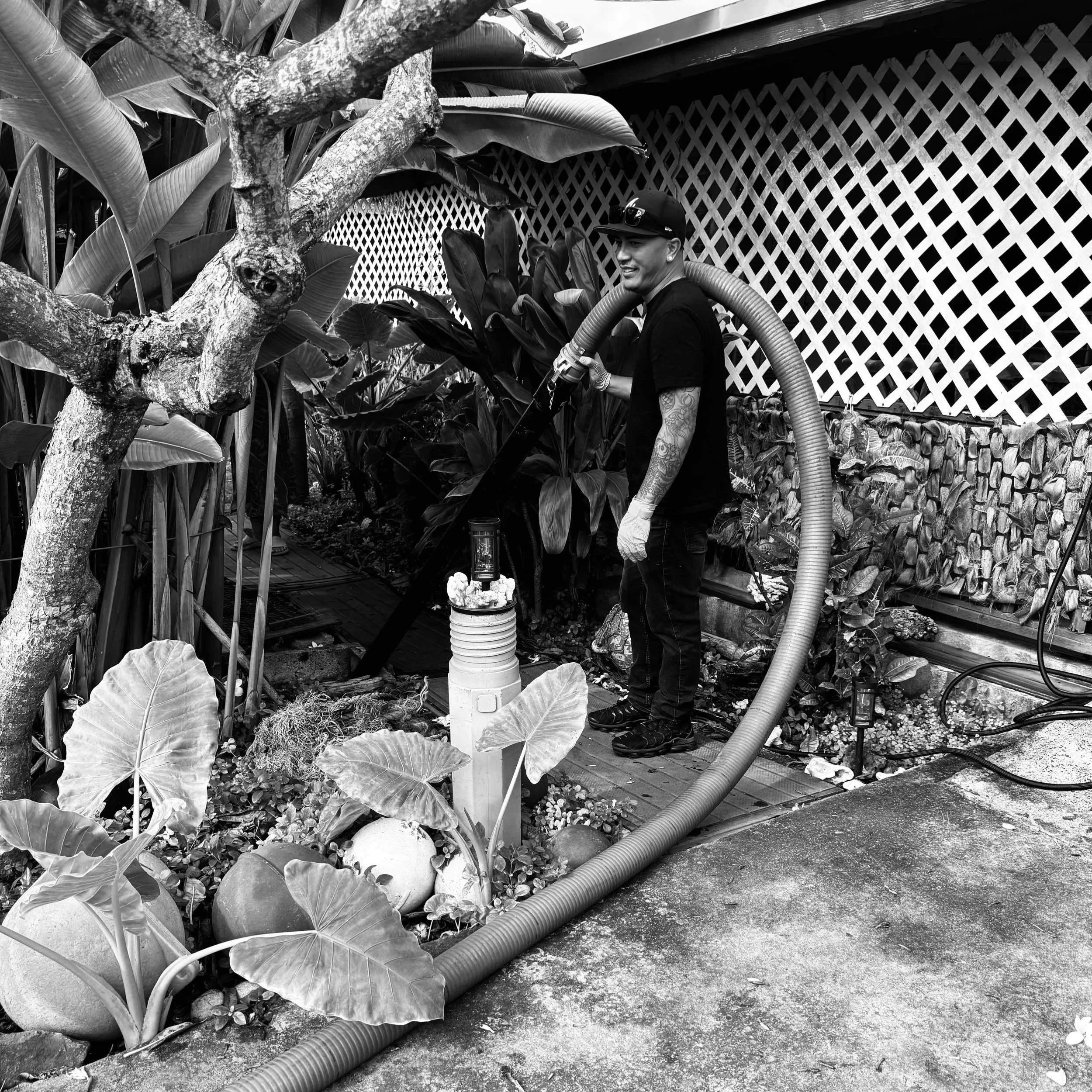 A man with tattoos and wearing a cap, gloves, and casual black clothing, is watering plants with a large hose in a garden with tropical plants and a lattice fence in the background.