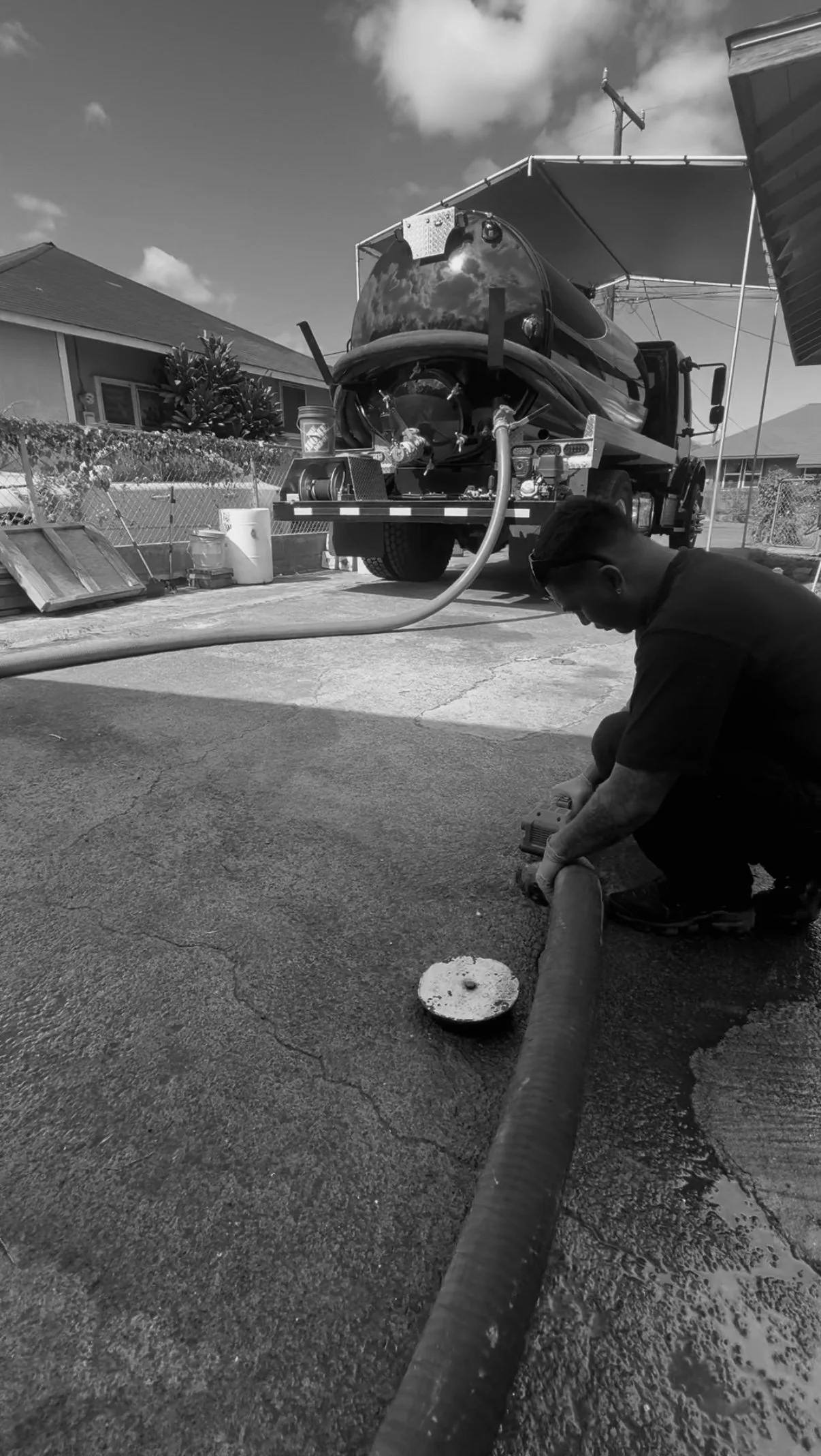 A person kneeling on a concrete driveway, working with a power tool attached to a large flexible hose connected to a water tanker truck, which is parked outdoors in a residential area on a sunny day.