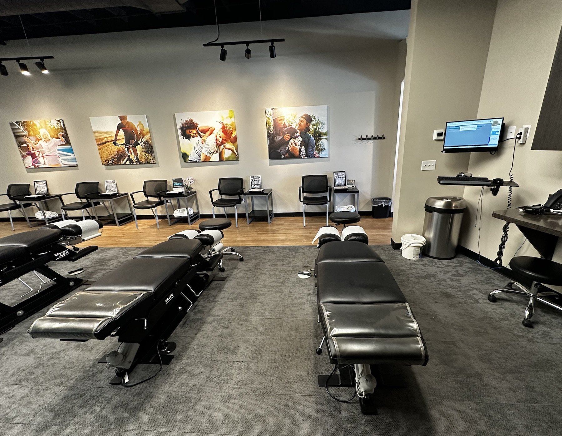 Medical examination room with two black therapy tables, a gray carpet, a row of chairs along the wall with photographs, and a computer monitor on the wall.