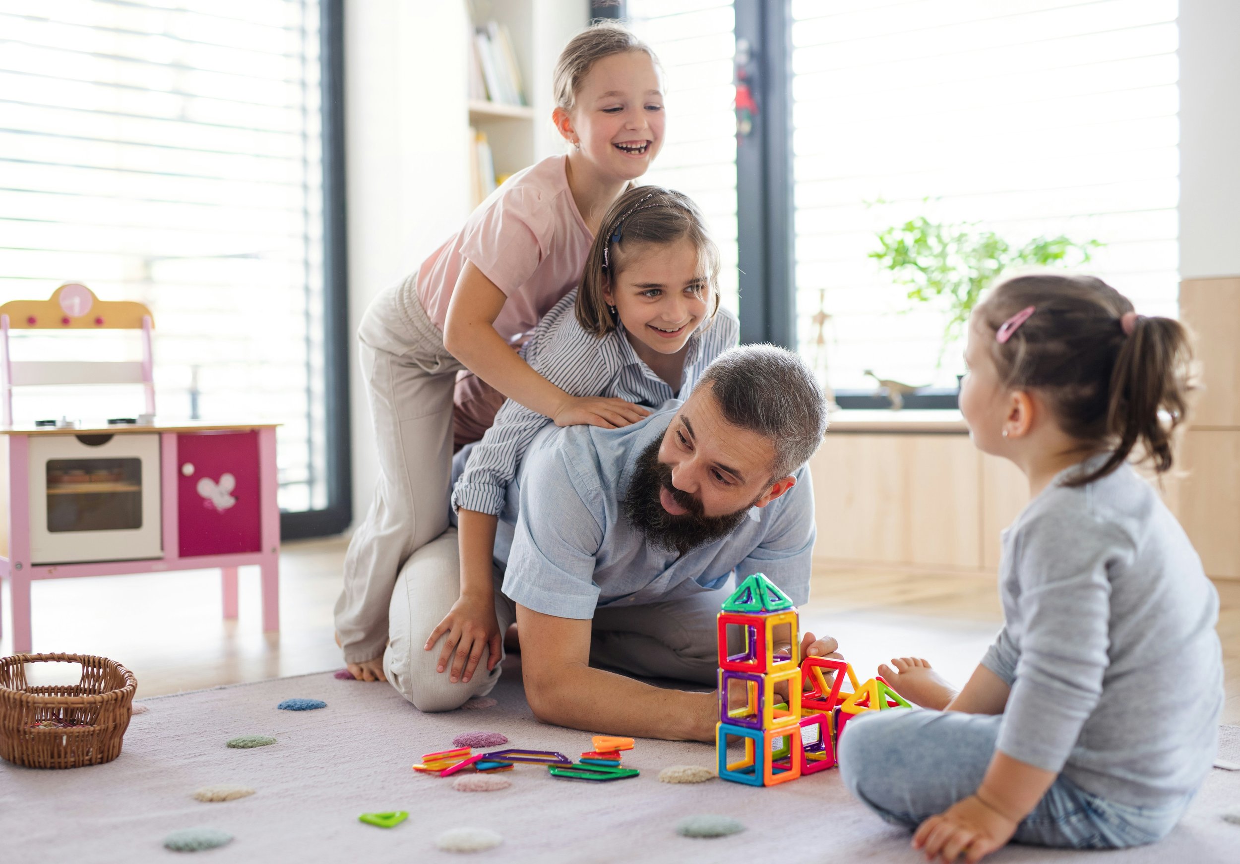 A group of four children and one adult playing with colorful building blocks on a beige carpet in a brightly lit room with large windows and a pink toy kitchen in the background.