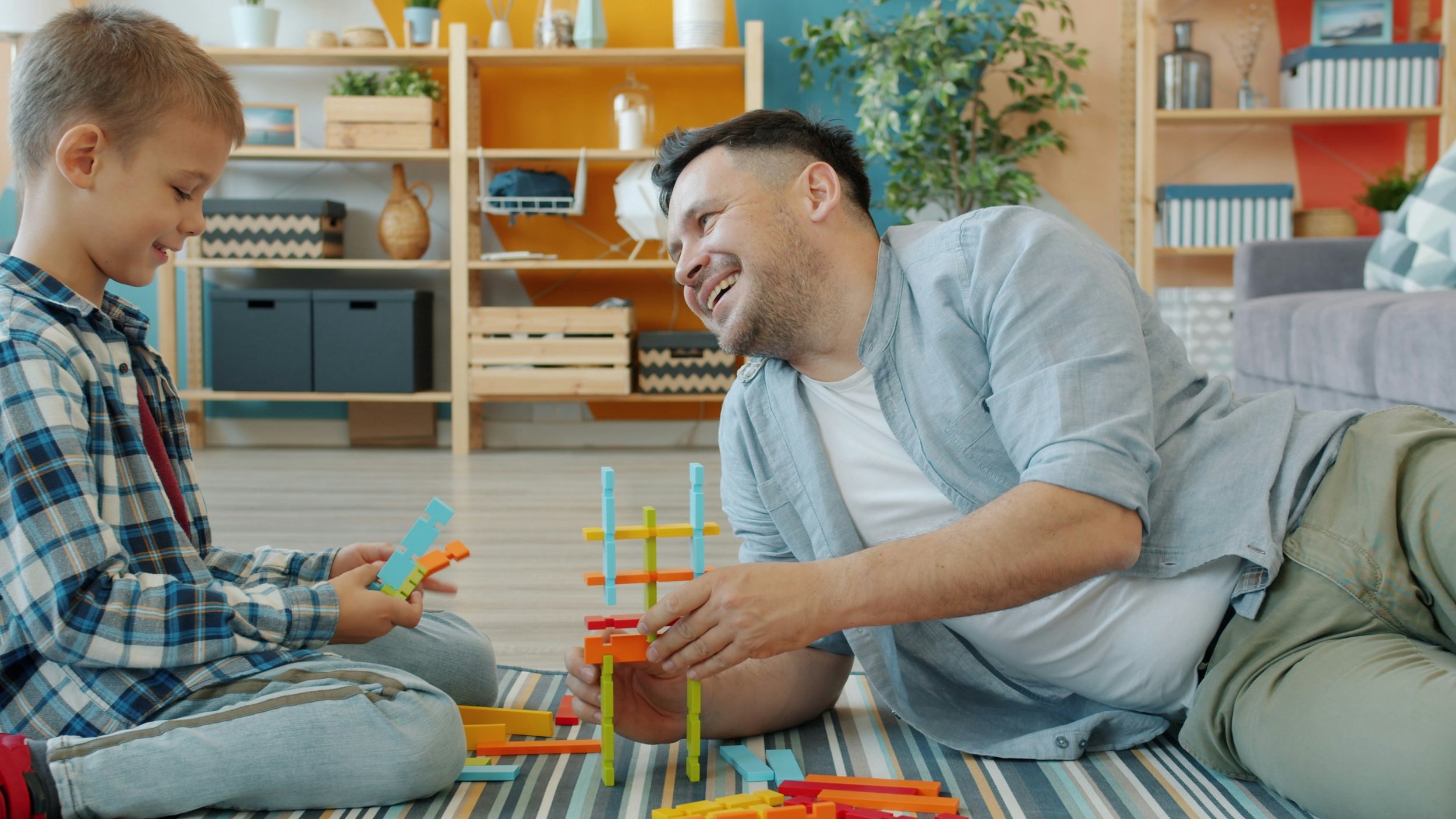 A man and a boy playing with colorful building blocks on a striped rug in a cozy living room.