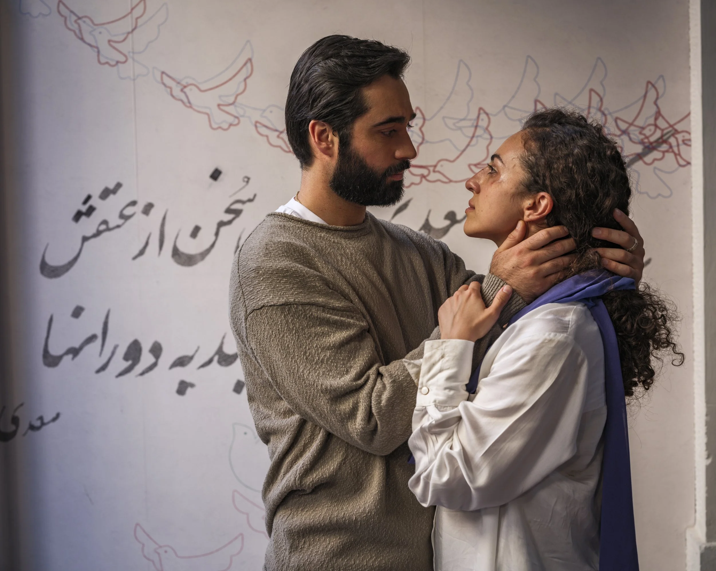 A man and woman are facing each other closely, holding each other's faces, in an emotional moment. Behind them, there is a wall with writing and drawings.